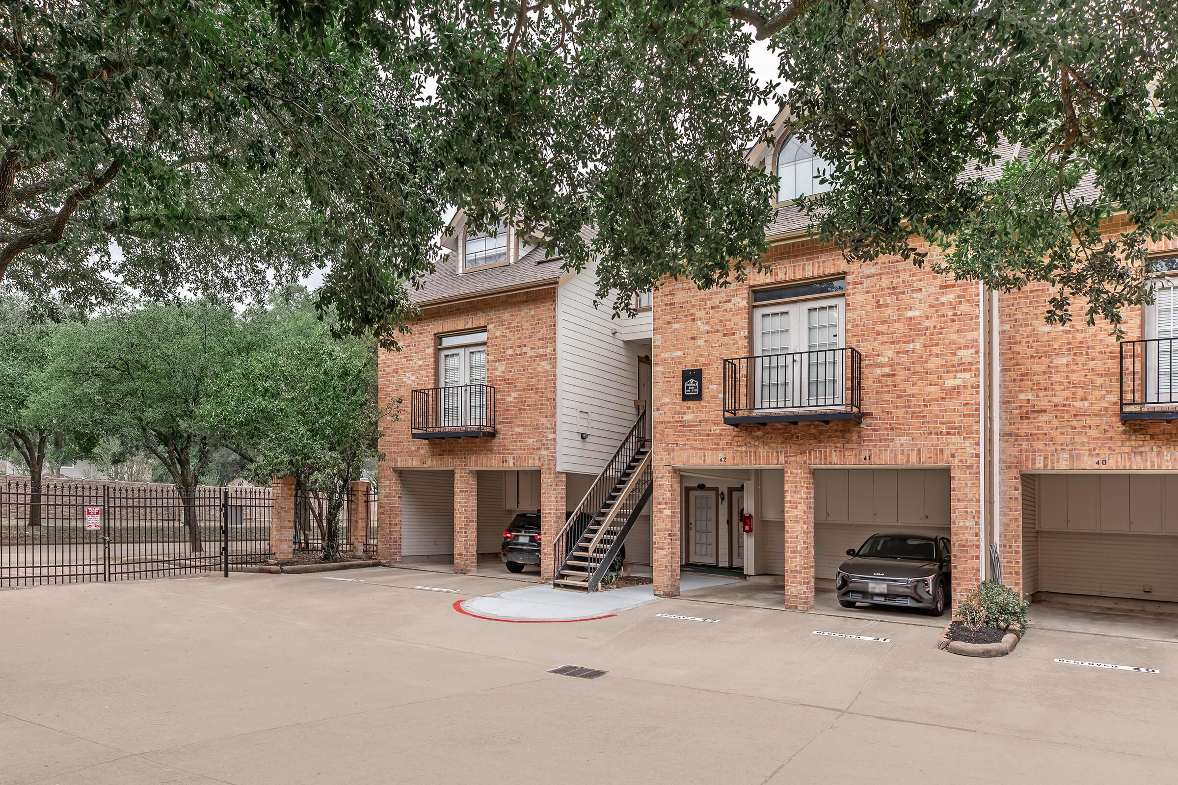 A view of a brick apartment complex featuring two units, each with a balcony. There are parked cars beneath the units, and a staircase leading to the upper level. Lush green trees surround the area, and a gated entrance is visible in the background. The parking area shows designated spaces.