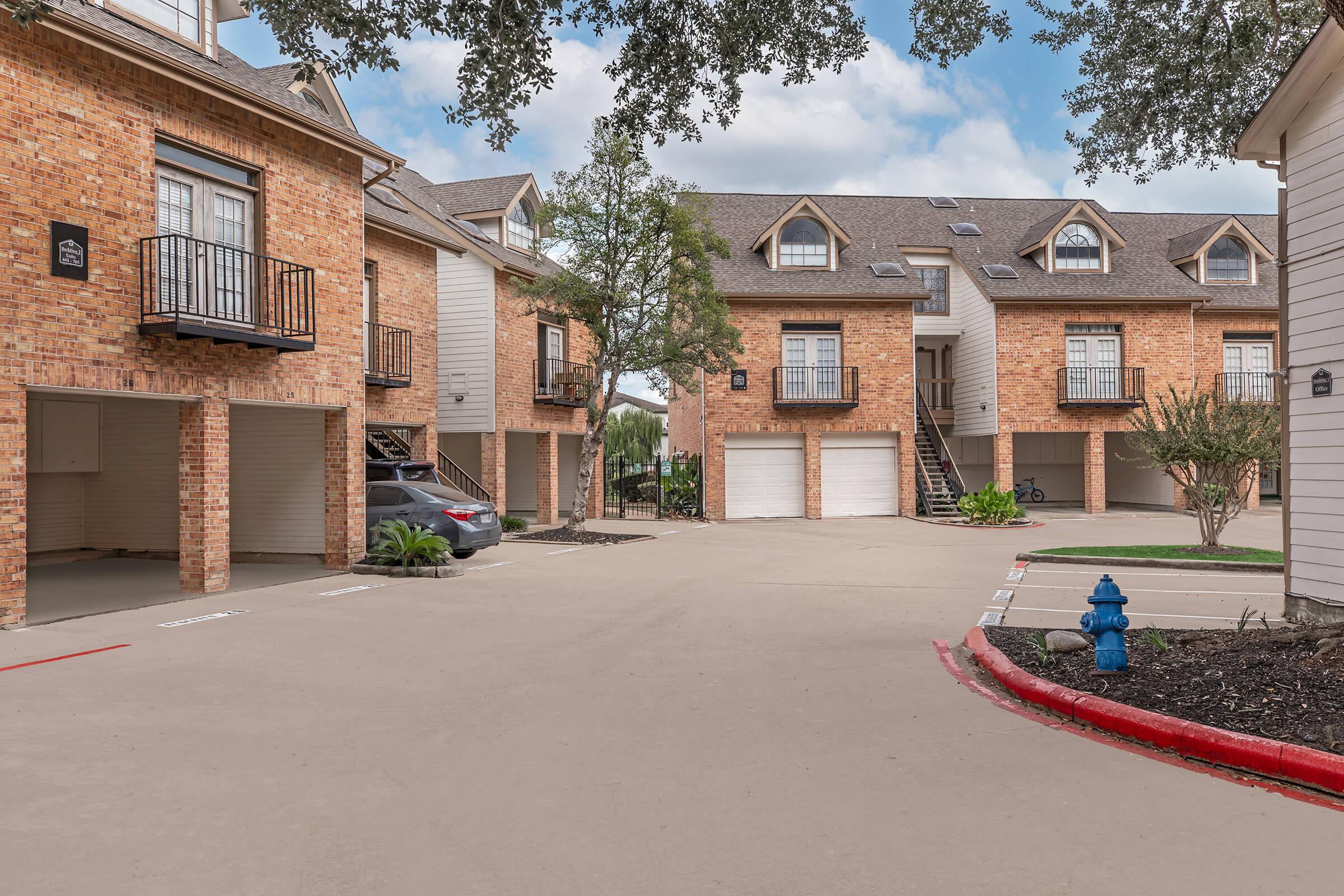 A view of a residential area featuring brick apartment buildings with balconies, garages underneath, and a paved parking lot. There are trees and landscaping, as well as a blue fire hydrant near the curb. The sky is partly cloudy, enhancing the appealing atmosphere of the community.