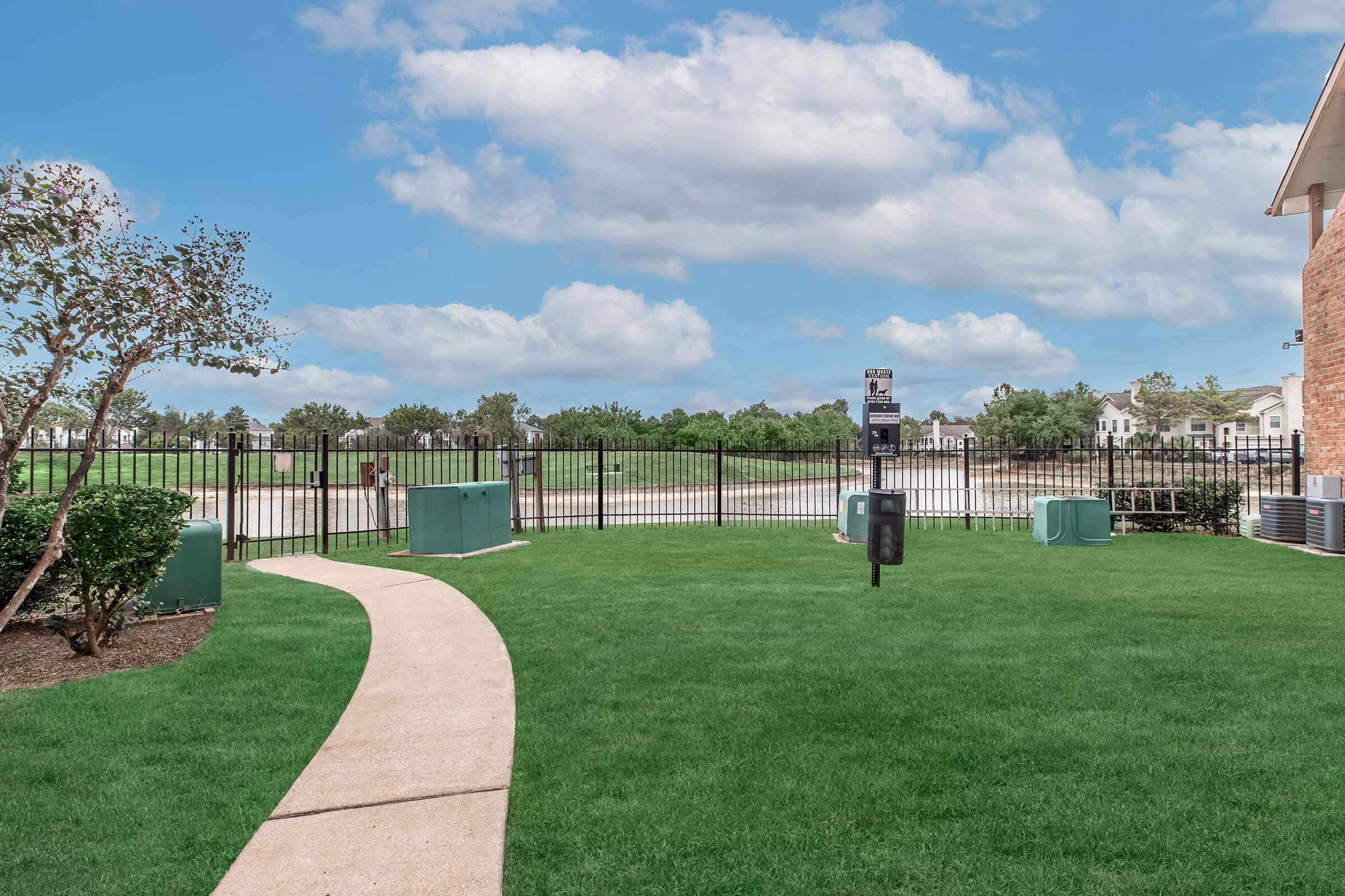A well-maintained grassy area with a paved pathway leading to a fenced waterway. There are utility boxes and picnic spots in the vicinity, surrounded by trees and bushes under a partly cloudy sky. The setting appears serene and welcoming, ideal for outdoor activities.