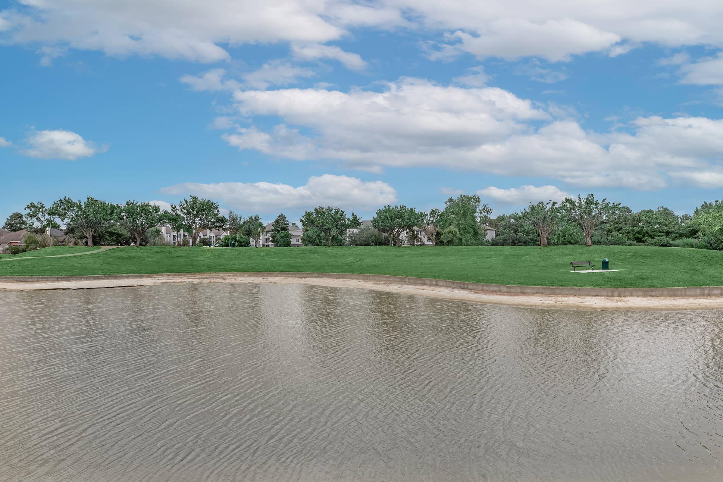 A serene landscape featuring a calm lake with a gentle shoreline, bordered by lush green grass. In the background, trees and residential buildings can be seen under a partly cloudy blue sky. A solitary bench is positioned near the water, inviting relaxation and reflection.