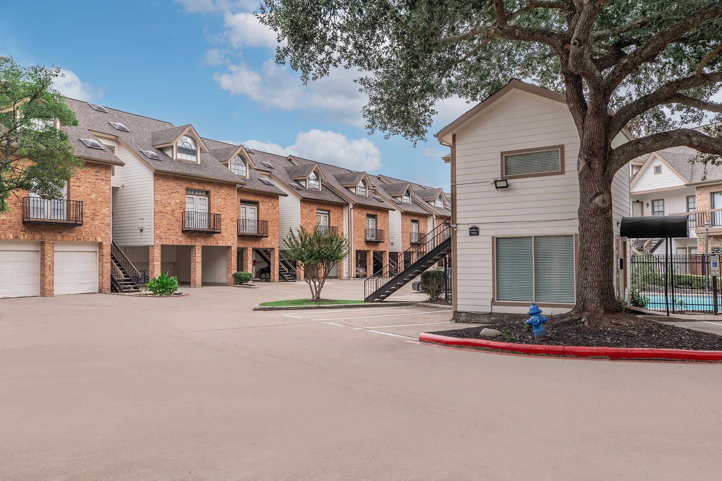 A view of a residential complex featuring two-story brick townhouses with balconies, adjacent to a white two-story building. The driveway is paved, lined with greenery, and a blue fire hydrant is visible. There is a swimming pool area in the background, surrounded by trees.