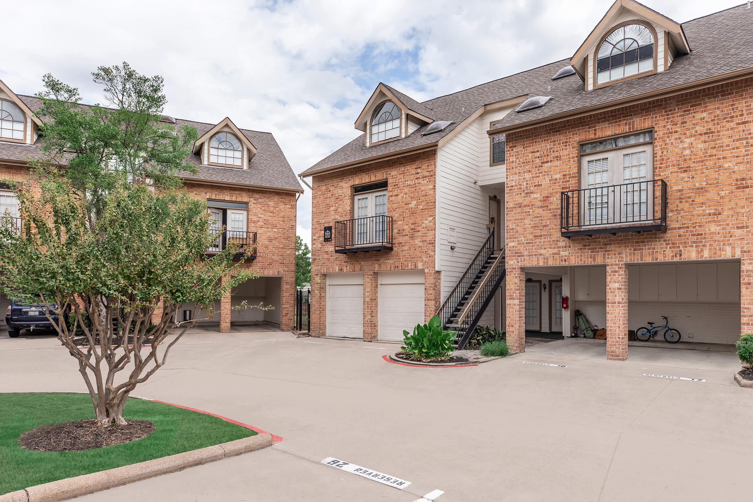 A well-maintained residential area with brick apartment buildings featuring balconies, a small courtyard with a tree, and parking spaces. The scene includes a bicycle parked near the entrance of one building, under a cloudy sky.