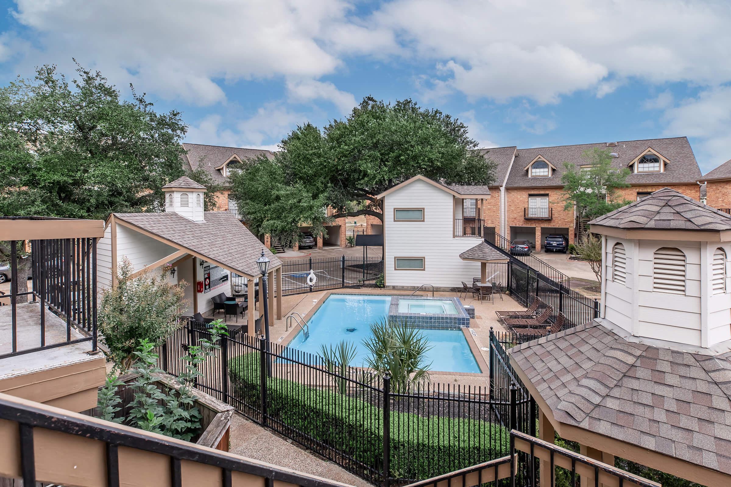A view of a residential area featuring a swimming pool surrounded by fencing. There are lounge chairs by the pool, a small gazebo, and several trees in the background. The setting includes two-story buildings with a well-maintained landscape, under a partly cloudy sky.