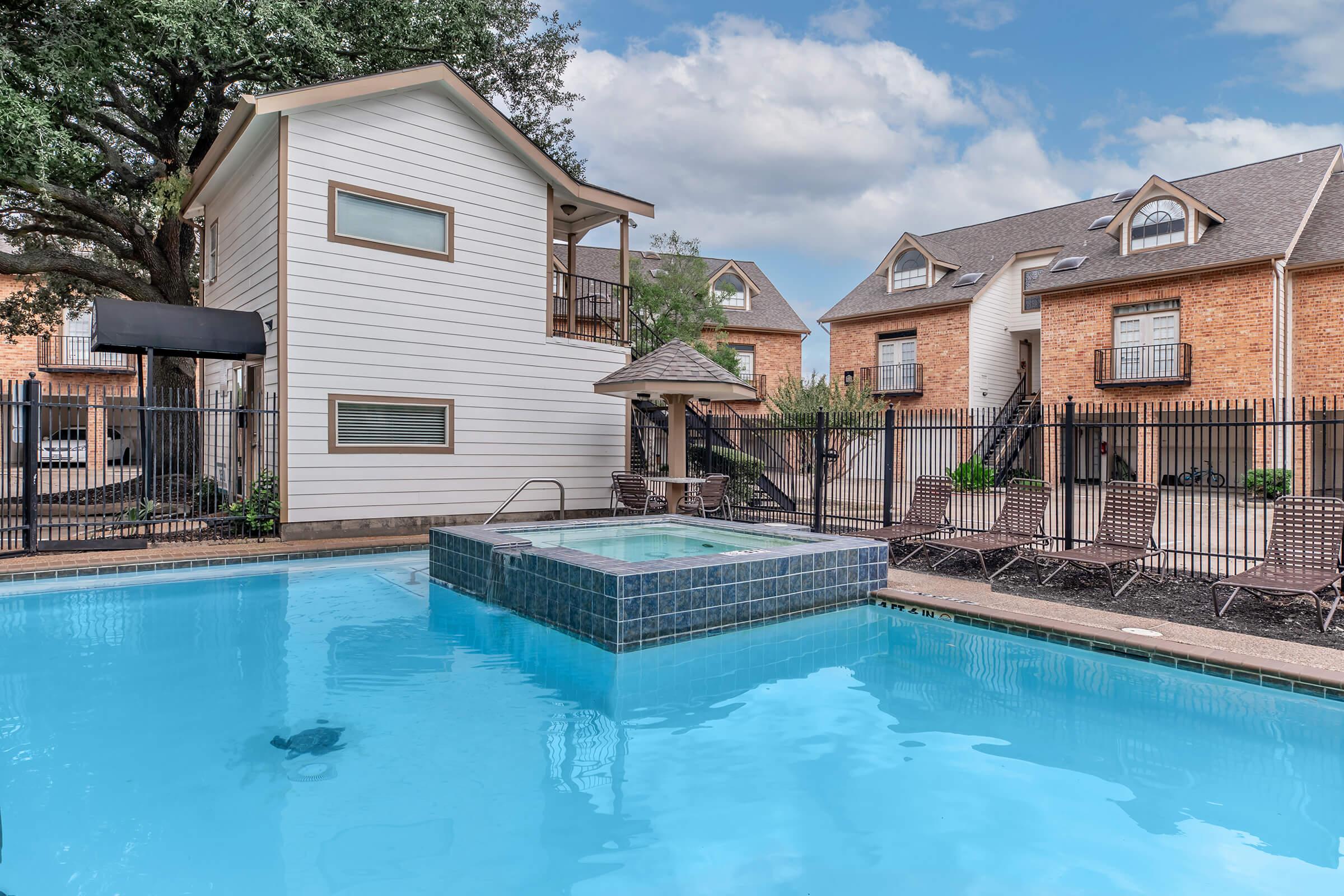 A clear blue swimming pool with a spa attached, surrounded by a black fence. There is a two-story light-colored building with a small gazebo nearby. In the background, there are two brick apartment buildings with balconies. The setting is sunny, with a few trees providing shade.