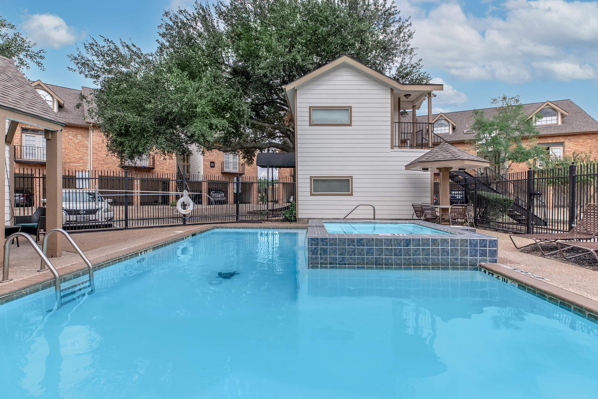 A bright outdoor pool area featuring a clear blue pool with a small hot tub nearby. Lush greenery surrounds the space, and a two-story building sits in the background. There are lounge chairs and a safety ring by the pool, creating a relaxing atmosphere in a residential community.