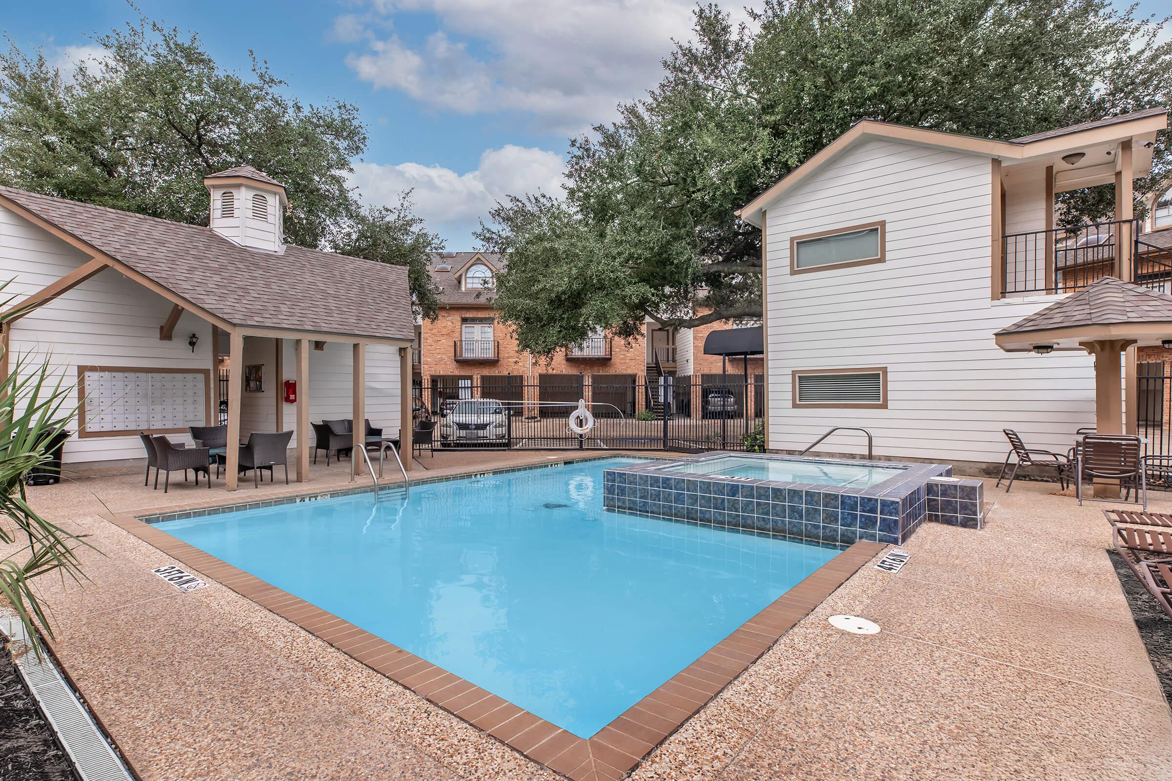 A clean swimming pool surrounded by a patio area, featuring a spa and several lounge chairs. Two residential buildings are in the background, with greenery and trees enhancing the outdoor space. The setting appears inviting and well-maintained.