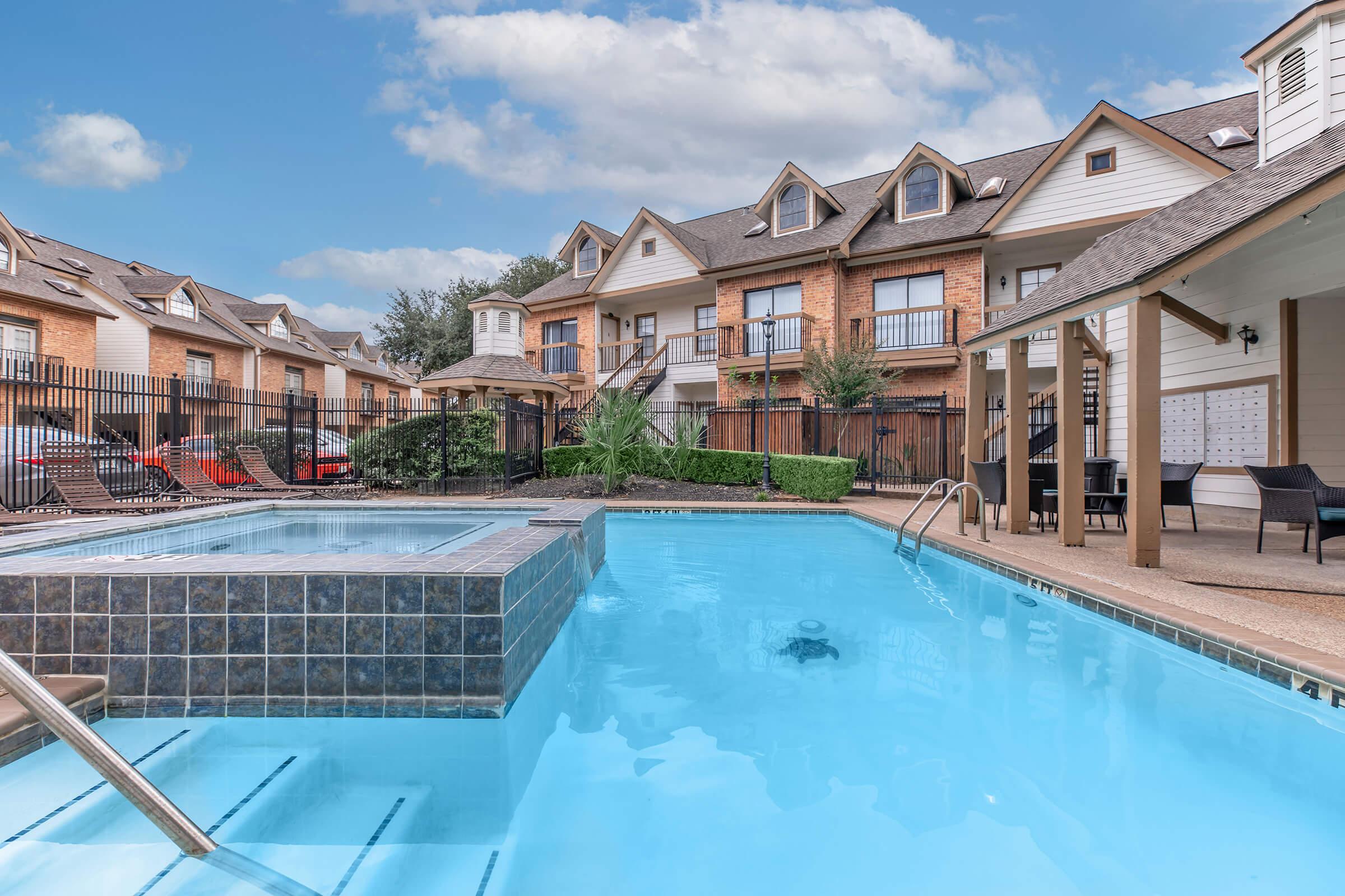A clean swimming pool with a hot tub on the side, set in an apartment complex. The pool area features lush landscaping and lounge chairs. In the background, well-maintained buildings with balconies are visible under a partly cloudy sky.