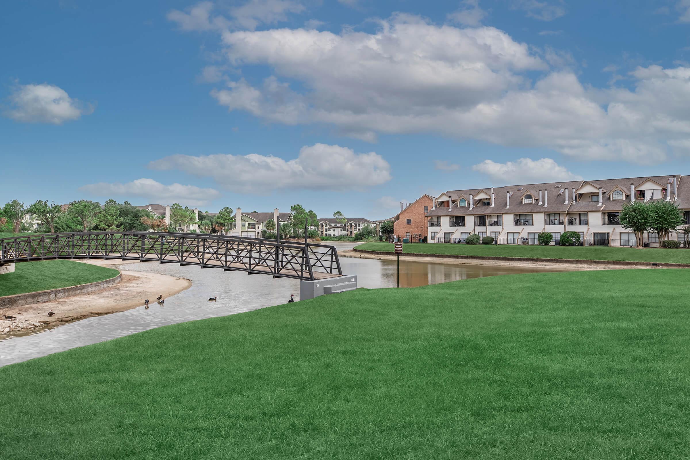 A serene landscape featuring a grassy area by a calm waterway, with a decorative wooden bridge arching over the water. On the right, there are modern apartment buildings surrounded by trees, under a partly cloudy blue sky. A few ducks are seen near the bank of the water.