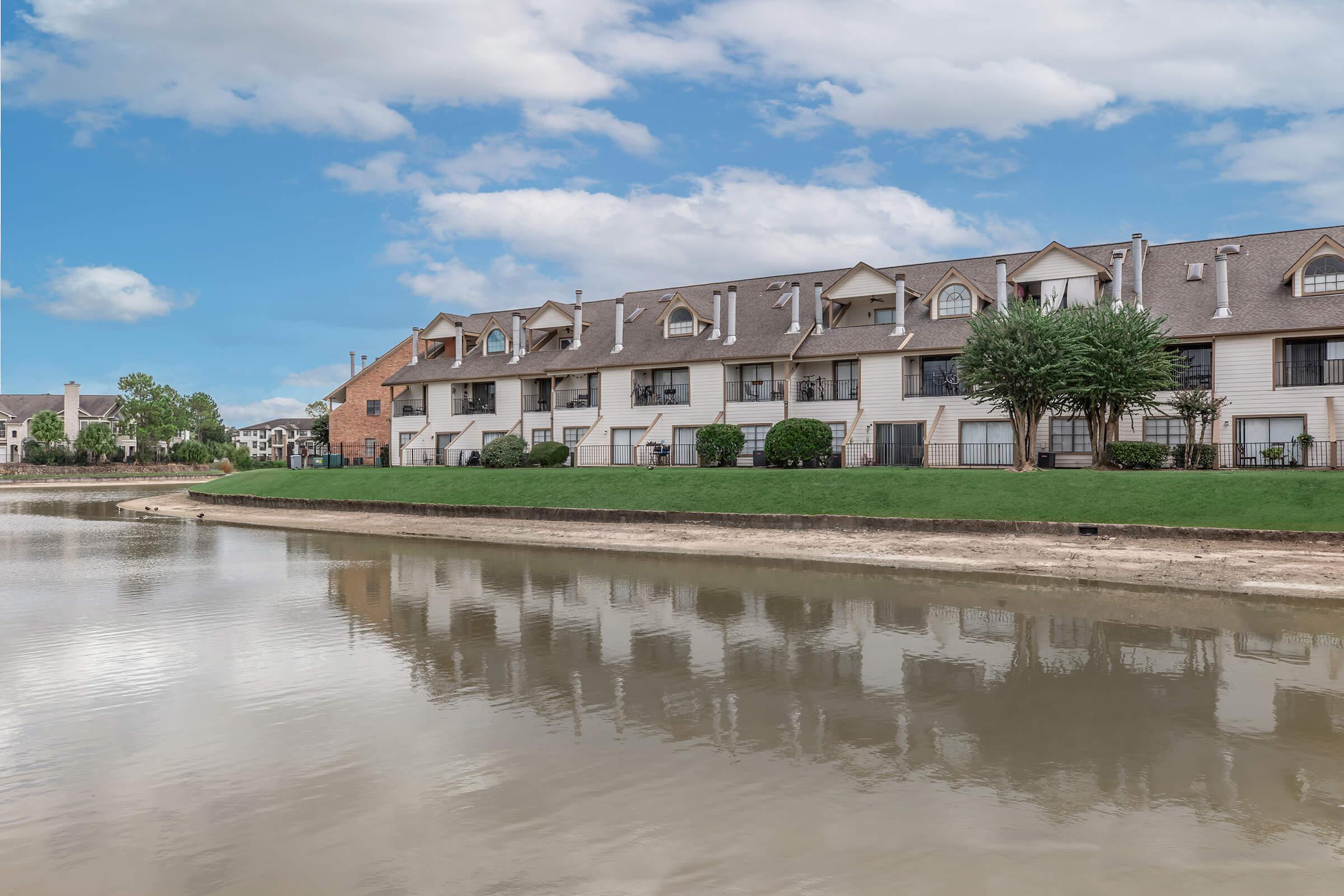 A view of a tranquil lake with a grassy shoreline, reflecting a row of residential buildings with a mix of brick and siding. The sky is partly cloudy, creating a serene and peaceful atmosphere. Lush green trees line the water's edge, enhancing the natural beauty of the scene.