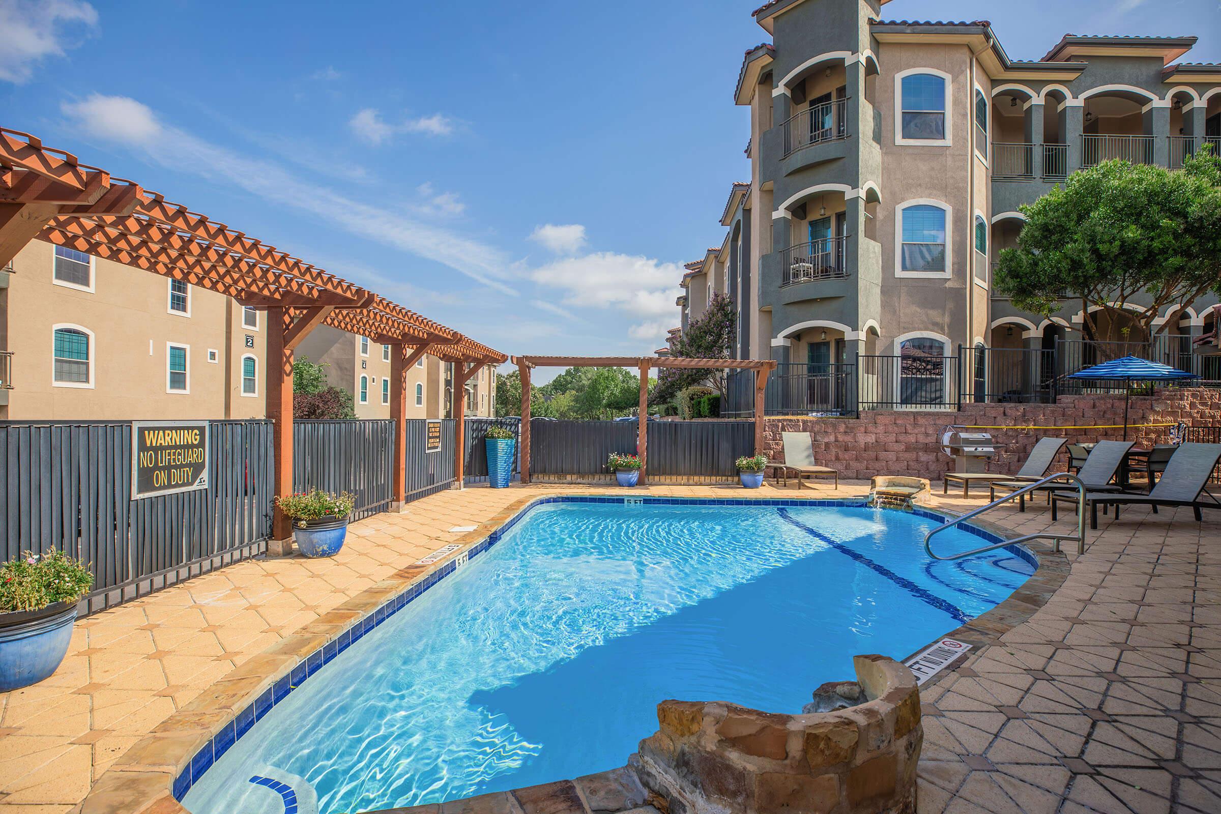 A bright, inviting pool area surrounded by apartment buildings, featuring a clear blue pool, lounge chairs, and potted plants. The pool deck has a stone border, and there's a warning sign visible on the fence. The sky is clear with a few clouds, creating a pleasant outdoor atmosphere.