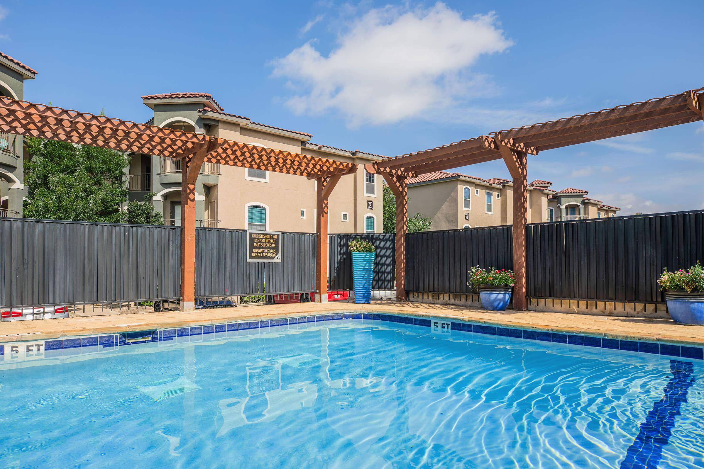 A clear blue swimming pool surrounded by wooden pergolas and potted plants. The pool area is enclosed by a dark fence, with apartment buildings visible in the background under a bright sky with a few clouds.