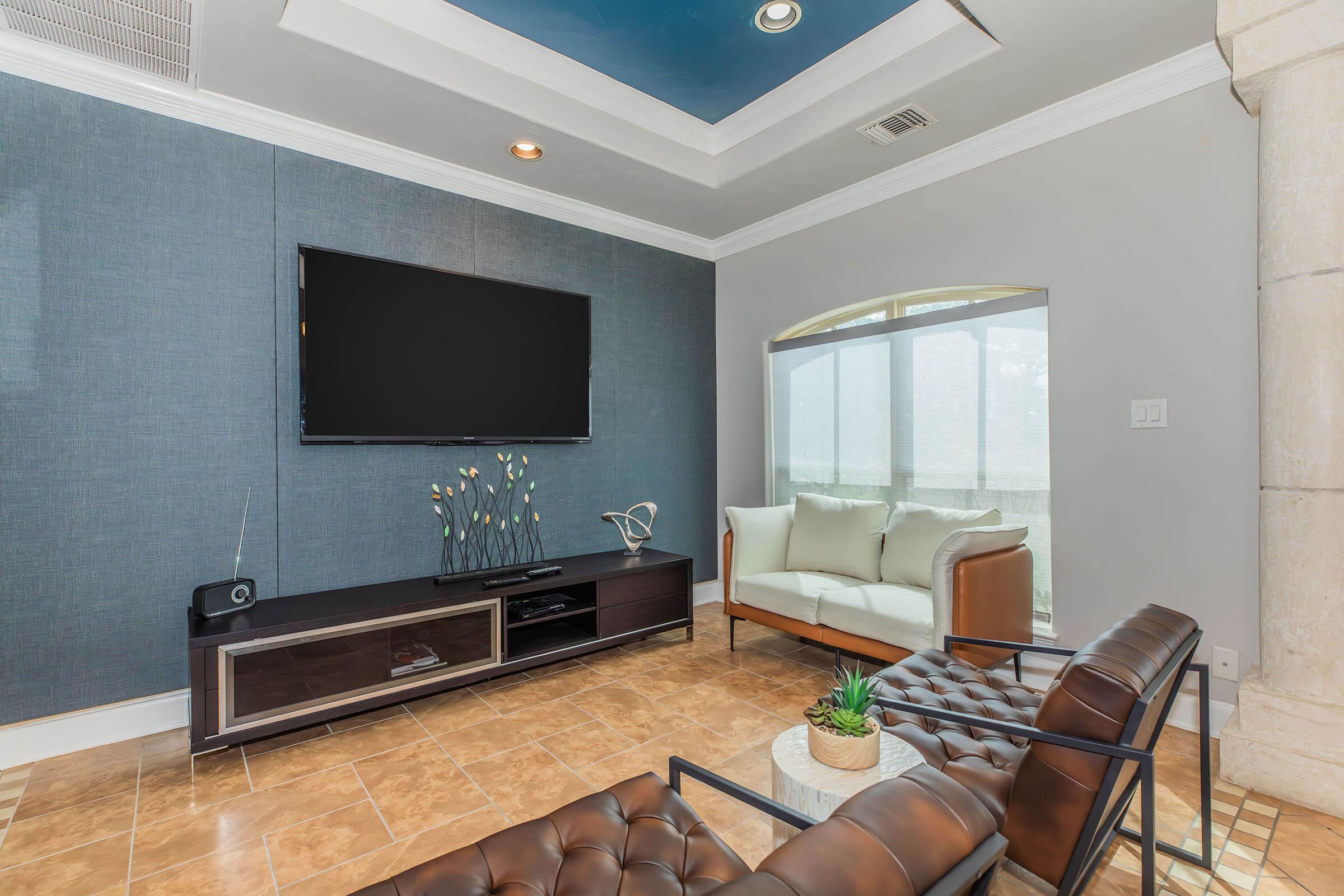 A modern living room featuring a flat-screen TV mounted on a blue accent wall, a sleek dark coffee table, and stylish brown chairs. The room has large windows allowing natural light, with a light-colored sofa and decorative plant on the table. The flooring is tiled in warm shades.