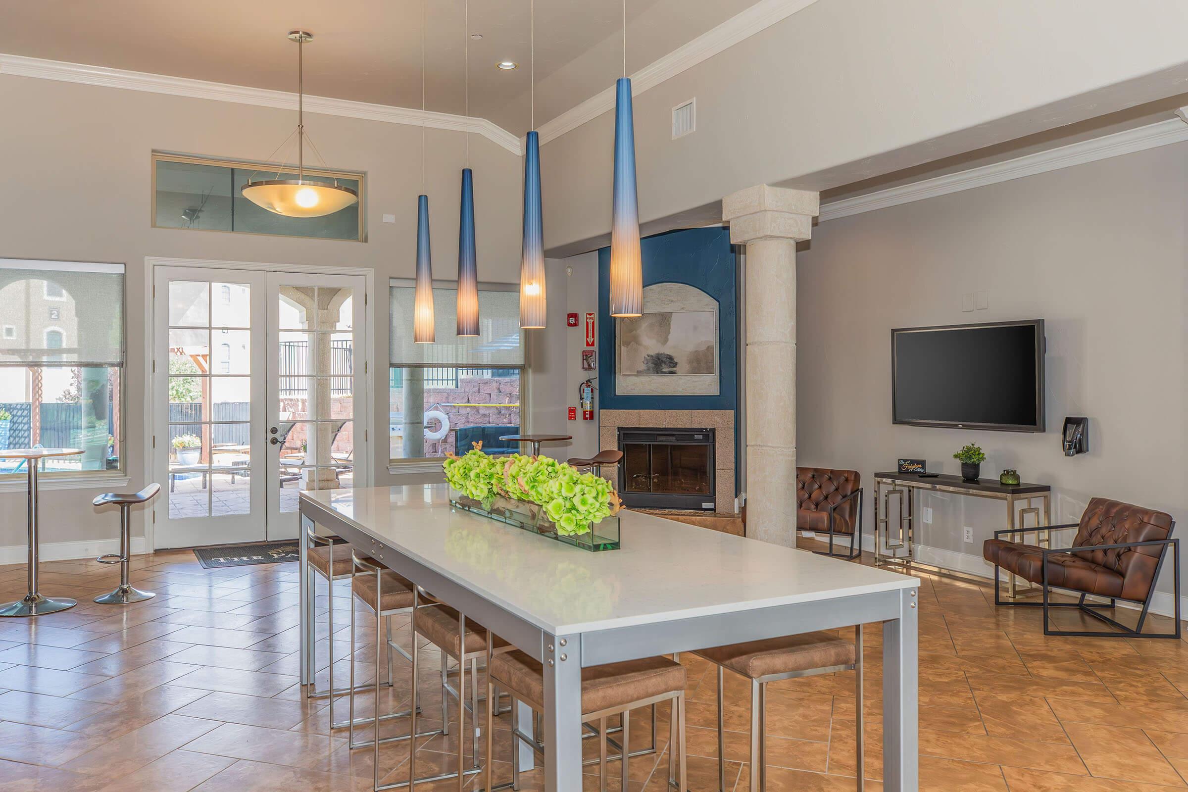 A modern dining area featuring a long table with decorative greenery, surrounded by stylish stools. Pendant lights hang above, illuminating the space. In the background, there is a large window with views of an outdoor pool area, and a cozy seating arrangement in a corner. A flat-screen TV is mounted on the wall.