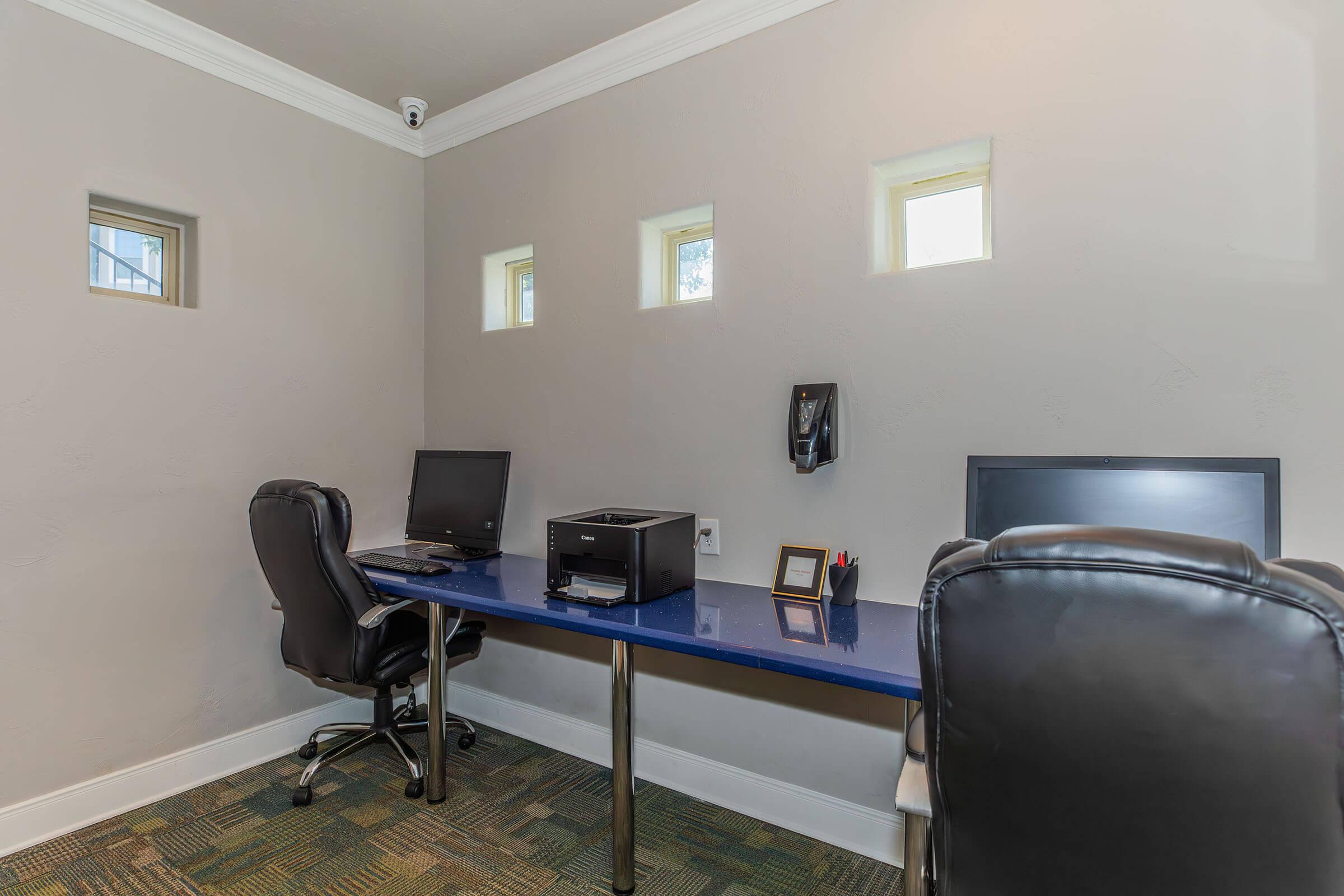 A minimalist office space featuring two black office chairs and a blue desk with two computer monitors. A printer is on the desk, along with a frame and a pen holder. The walls are light gray, and there are small rectangular windows letting in natural light. A security camera is mounted on the ceiling.