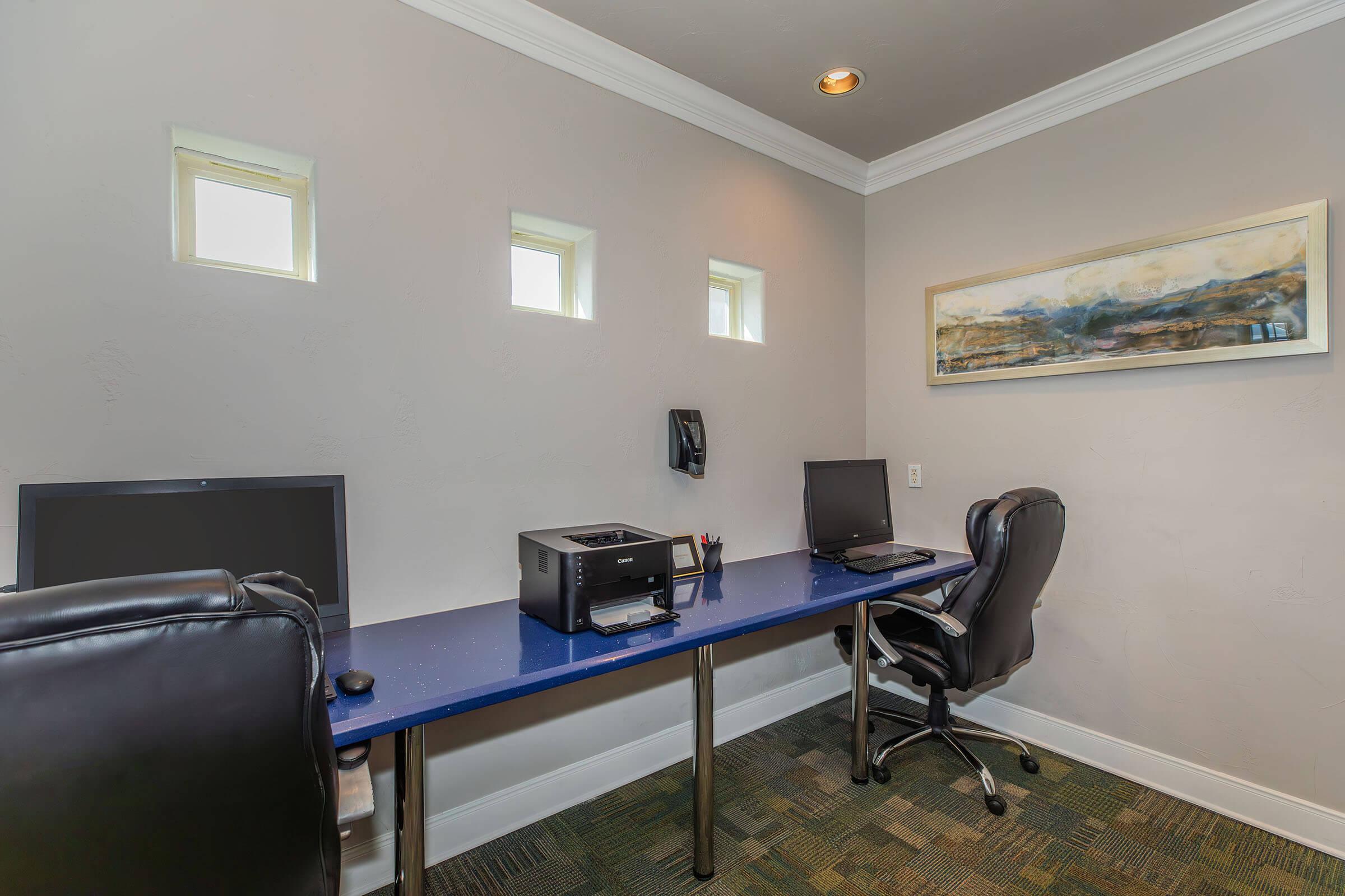 A modern office space featuring a long blue desk with two computer monitors and a printer. There are two black office chairs, and the walls are light gray with three small windows. A framed artwork hangs on the wall, adding a touch of decoration to the minimalist environment.