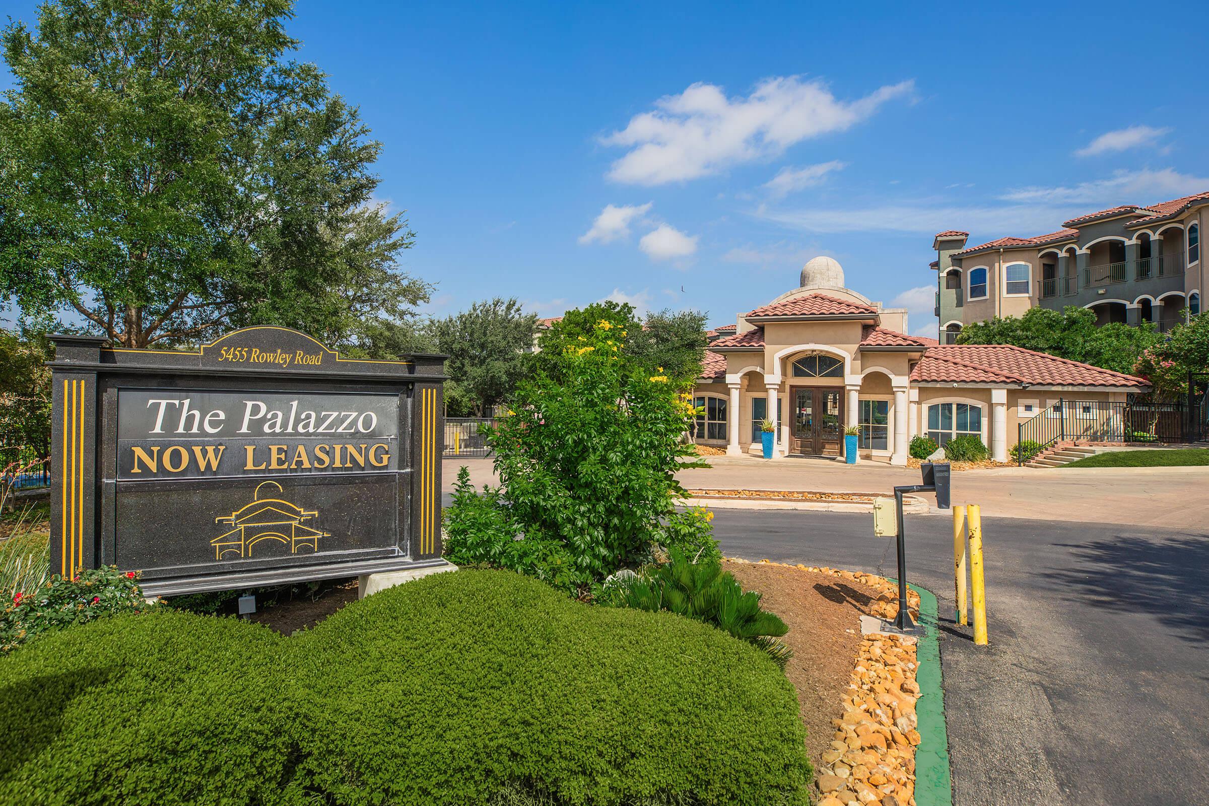 A well-maintained apartment complex sign reading "The Palazzо Now Leasing" is prominently displayed in front of the property. The entrance features a stylish building with a dome and landscaped greenery. Clear blue skies provide a bright atmosphere, indicating a welcoming residential area.