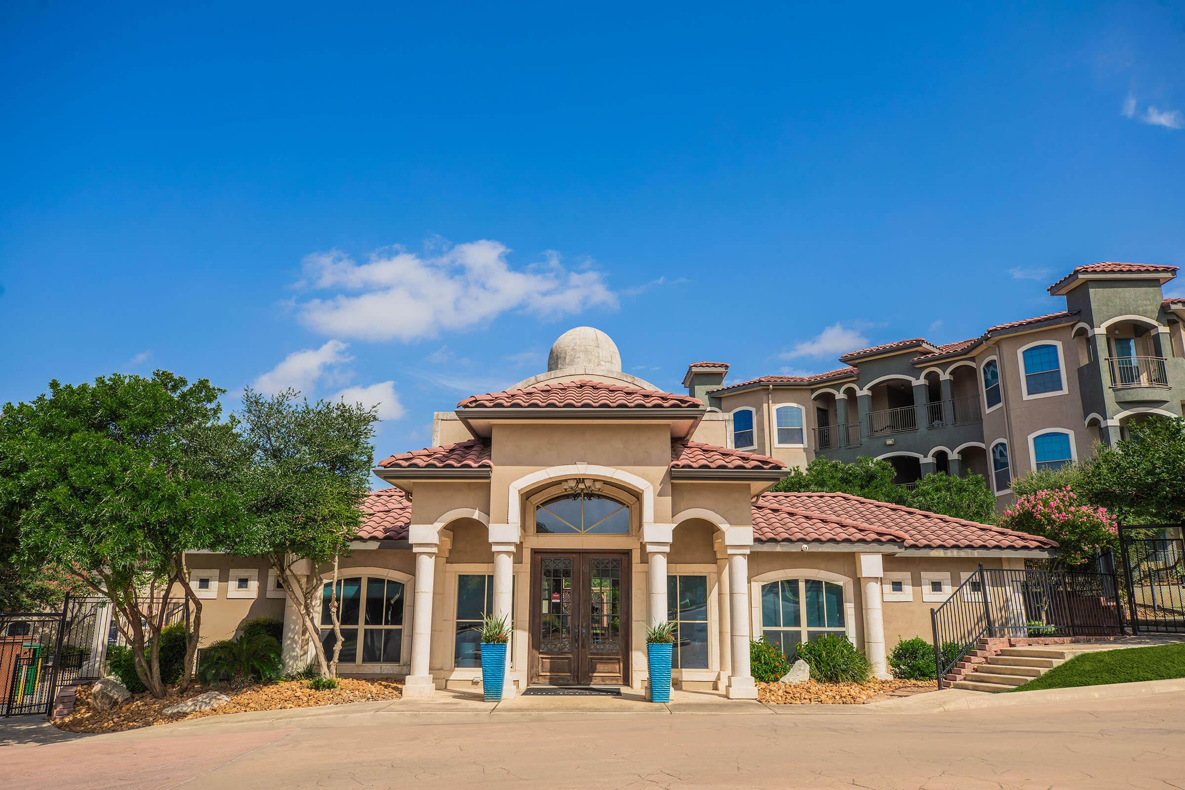 A picturesque building with a terracotta-tiled roof and arched entrance, surrounded by greenery. The facade features stone finishes and large windows, while a clear blue sky completes the scene. The background includes a multi-story residential complex with similar architectural style.