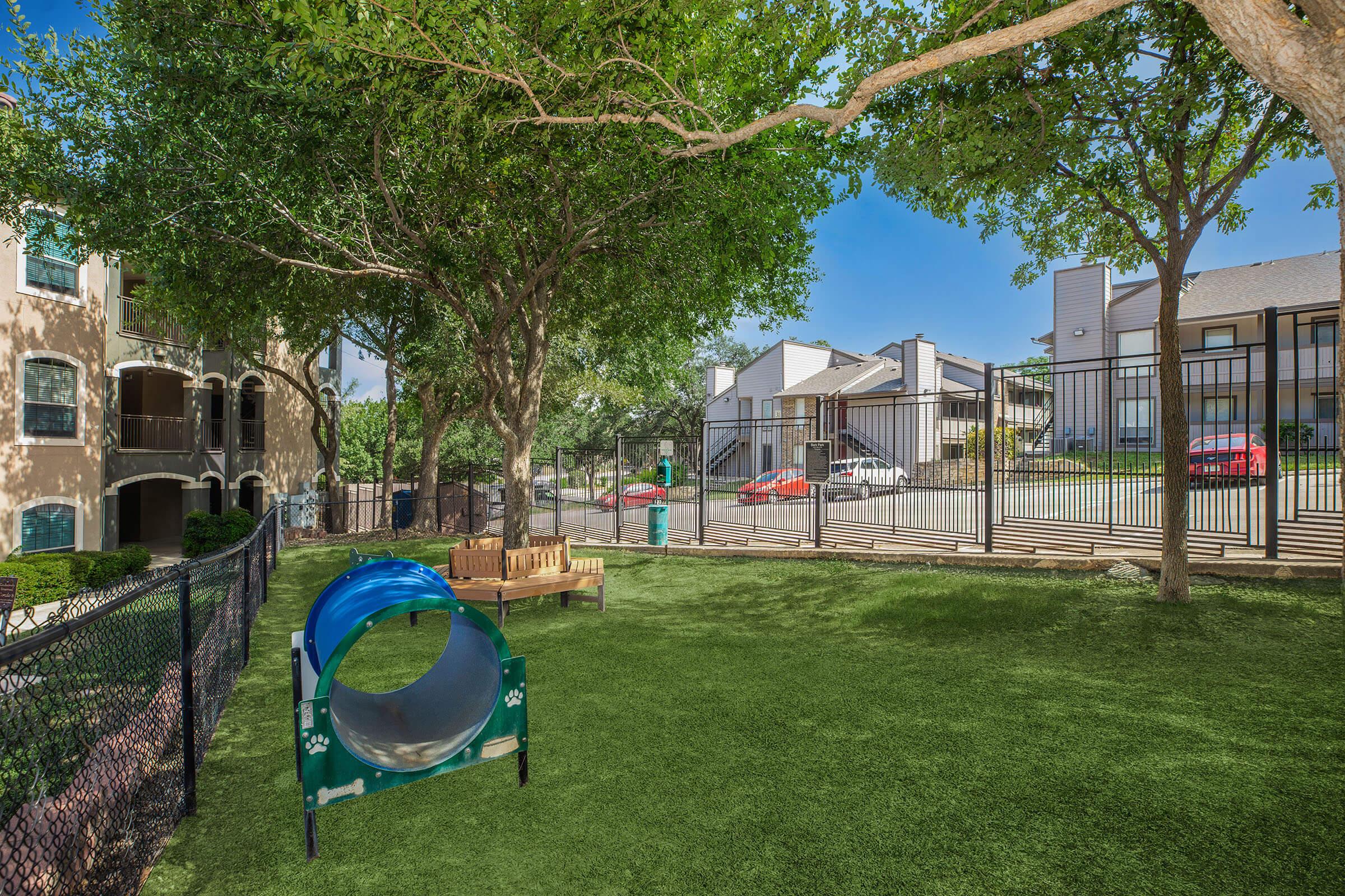 A grassy play area featuring a blue slide, benches, and surrounded by trees. The background shows a gated community with apartment buildings and parked cars, creating a serene outdoor space for relaxation and play.