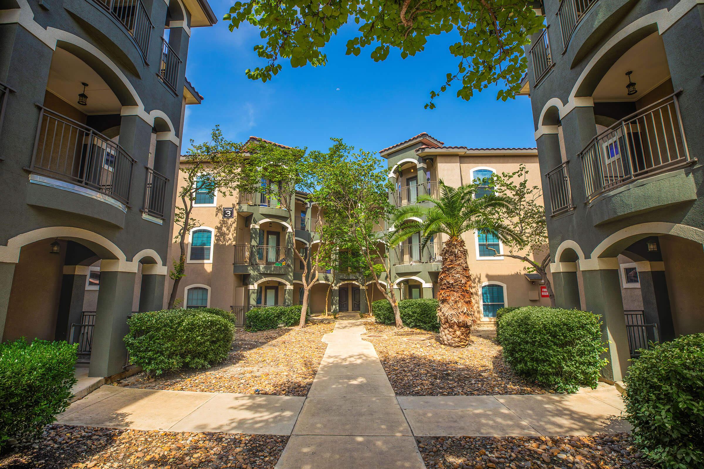 A view of a landscaped courtyard surrounded by modern apartment buildings. The path through the courtyard is lined with smooth stones and greenery, featuring palm trees and shrubs. The clear blue sky provides a bright backdrop for the pleasant outdoor space.