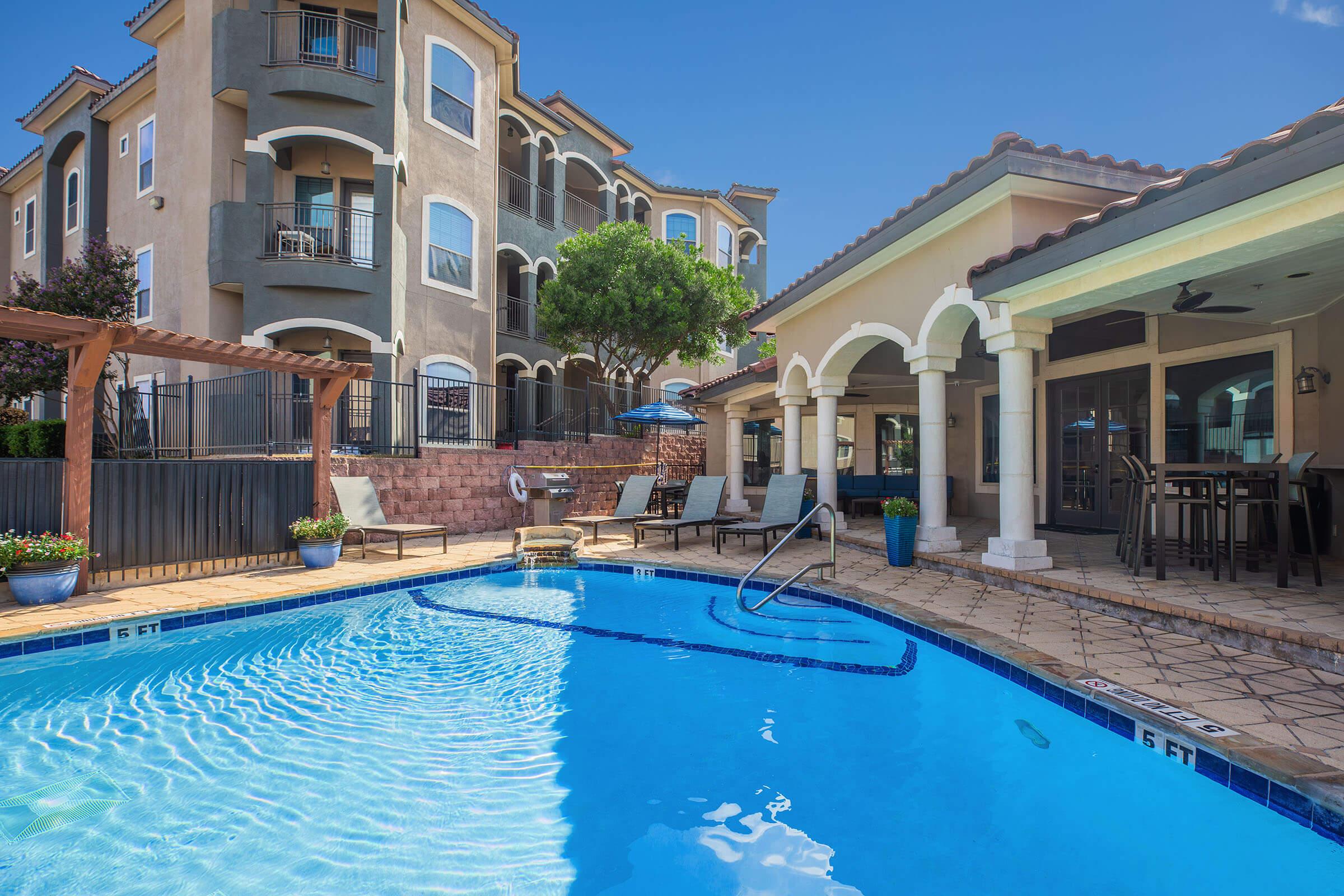 A bright and inviting outdoor pool area featuring crystal clear water, surrounded by decorative tiles. Nearby, there are comfortable seating arrangements under shaded patios, lush greenery, and a backdrop of modern apartment buildings against a clear blue sky.