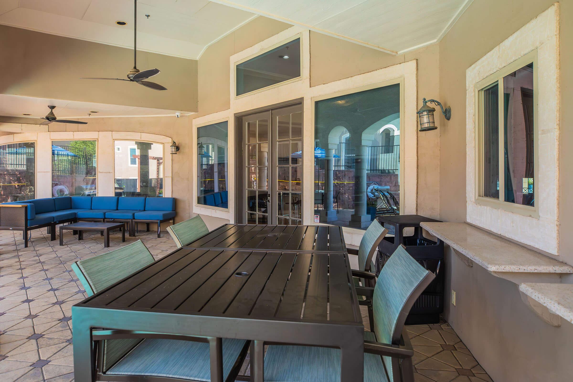 A spacious outdoor patio area featuring a large table with several chairs, a blue sectional sofa, and large windows that provide a view of an outdoor space. Ceiling fans are mounted above, and there is a light fixture attached to the wall, creating a comfortable and inviting atmosphere.