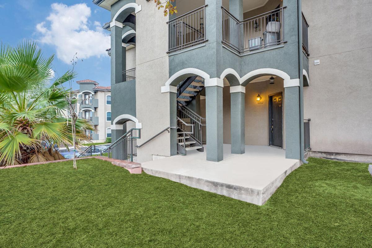 A view of a modern apartment exterior featuring a staircase leading to an entrance, with a well-maintained lawn area. The building has arched architectural details and a tropical palm tree nearby, under a partly cloudy sky.