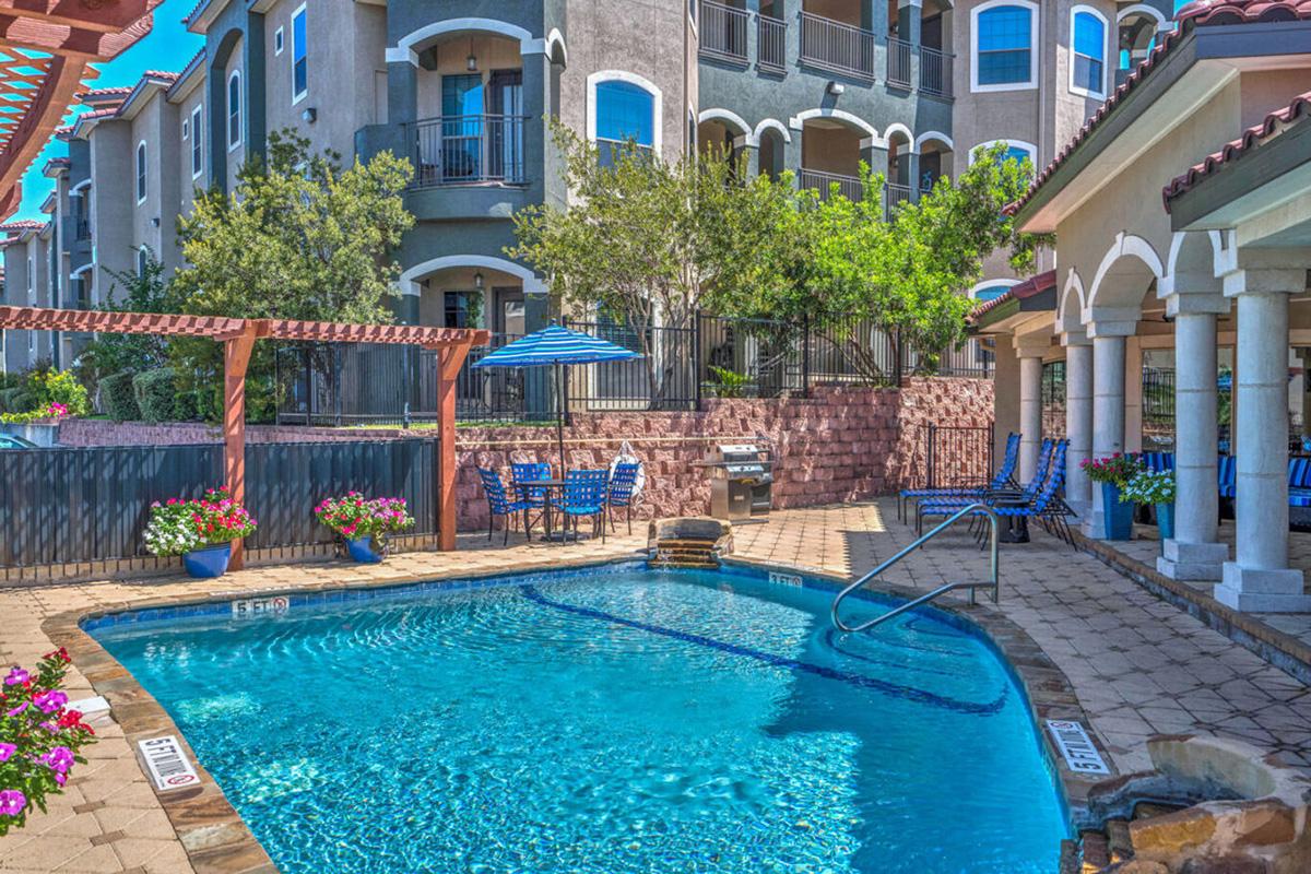 A vibrant pool area surrounded by elegant apartment buildings. The scene includes a blue swimming pool, lounge chairs, and shaded seating under a gazebo, with colorful flowers enhancing the landscape. The bright blue sky adds to the inviting atmosphere of this outdoor space.