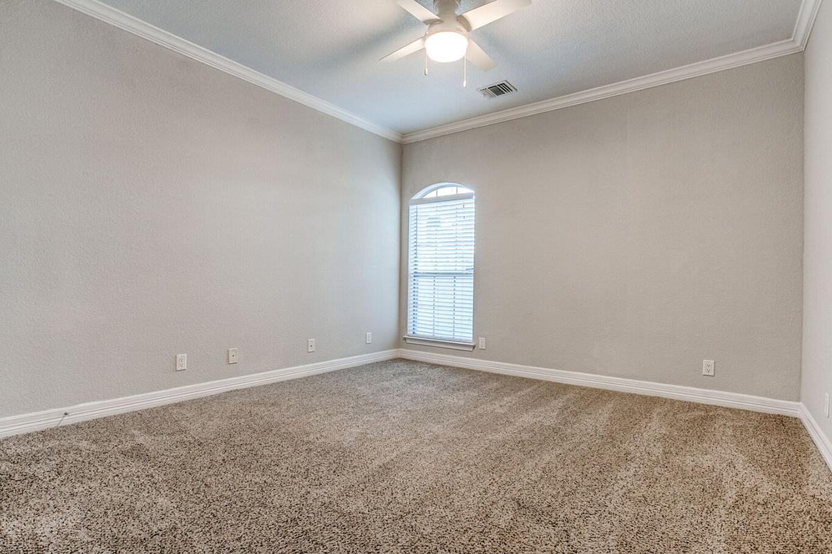 A spacious, empty room featuring light gray walls, a ceiling fan, and a window with white blinds. The floor is covered in plush beige carpet, and there is a decorative arched window near the ceiling. The overall ambiance is bright and inviting, suitable for various uses.