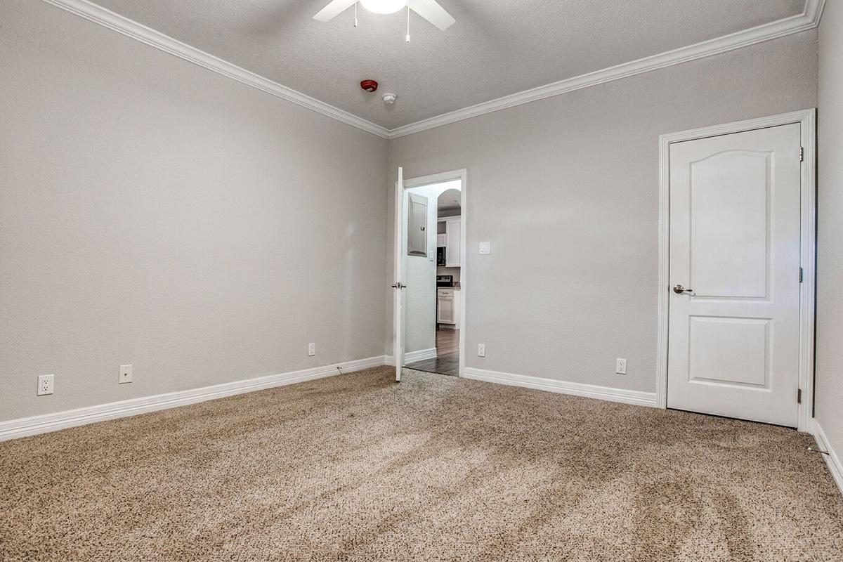 Empty room with light gray walls and carpeted flooring. A ceiling fan is visible, and a white door is slightly ajar, leading to another room. The space is well-lit and clean, with baseboards and a smoke detector on the ceiling. The overall atmosphere is bright and minimalistic.