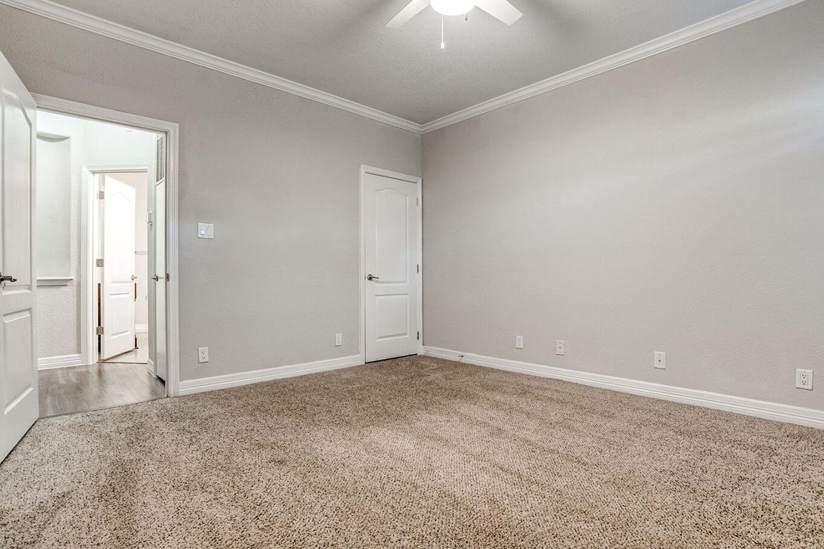 A sparsely furnished room featuring beige carpet and light gray walls. There is a single ceiling fan and a door leading to another room. The room appears bright and well-maintained, with no furniture or decorations present.