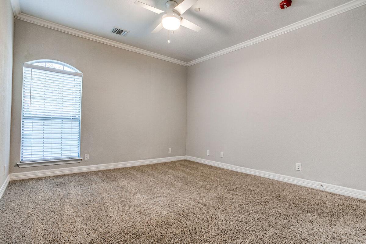 A spacious, empty room featuring light-colored walls, a light fixture on the ceiling, and a large window with white blinds. The floor is covered in tan carpet, creating a cozy atmosphere. The room has decorative crown molding and is well-lit with natural light coming from the window.