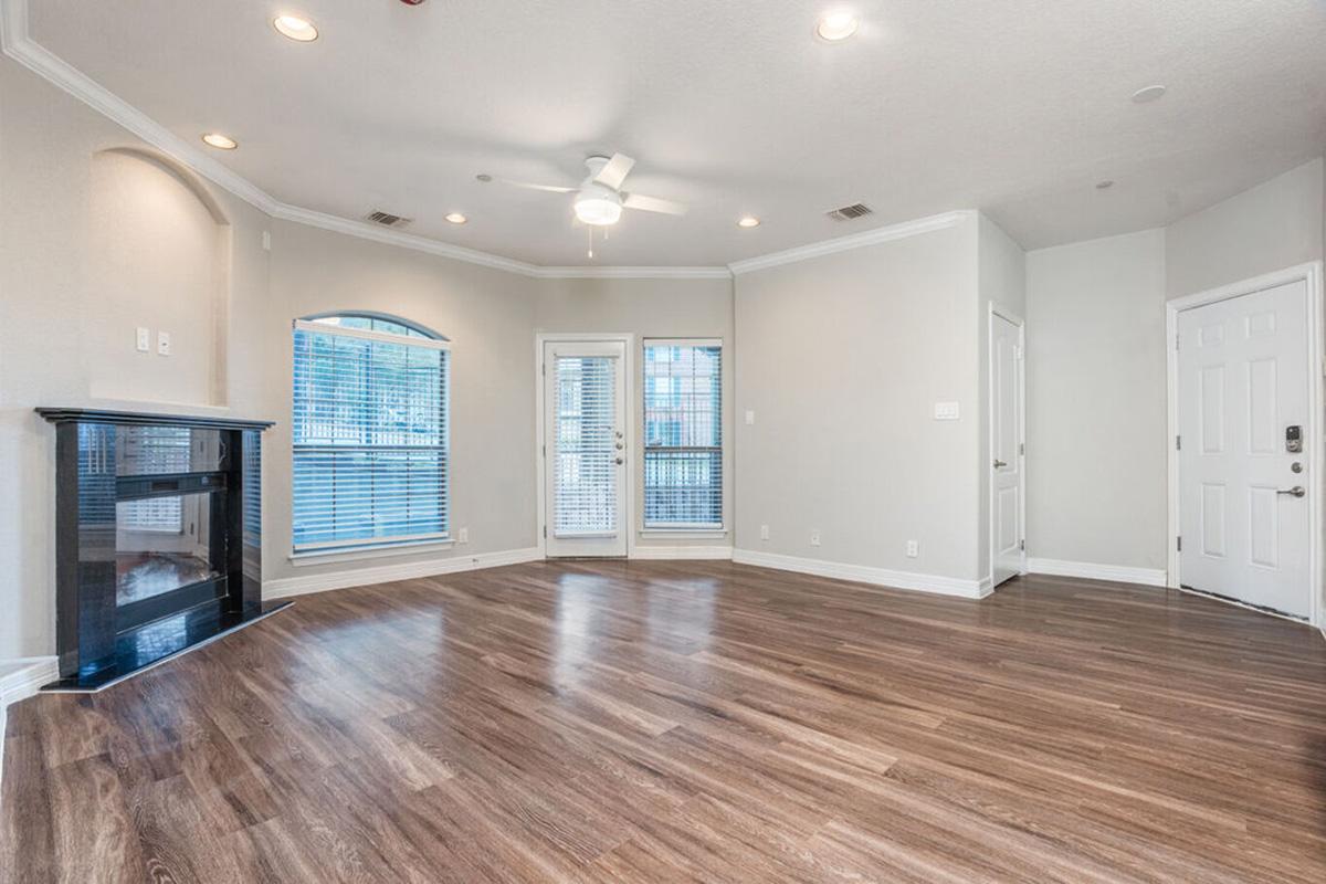A spacious living room with hardwood floors, featuring a ceiling fan, large windows with blinds, and a fireplace on the left side. There's an entryway on the right with a door leading outside, creating a bright and inviting atmosphere. The walls are painted in a light, neutral color.