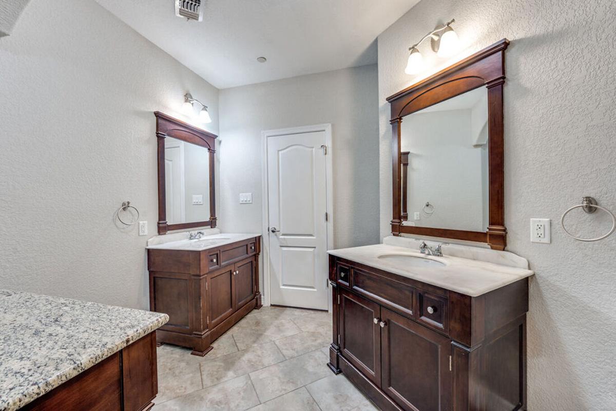 A modern bathroom featuring dual wooden vanities with marble countertops and matching mirrors. The walls are painted in a light gray, and the floor is tiled. Two wall-mounted lights illuminate the space, creating a clean and bright atmosphere. A white door is visible in the background.