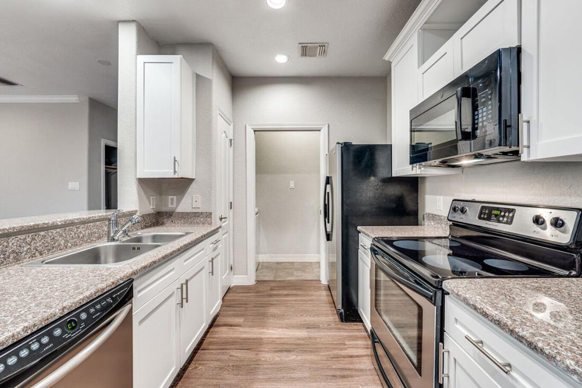 Modern kitchen featuring white cabinets, granite countertops, stainless steel appliances including an oven, microwave, and dishwasher, and a dark refrigerator. The space has wood-style flooring and a doorway leading to another room in the background. Brightly lit with recessed lighting.