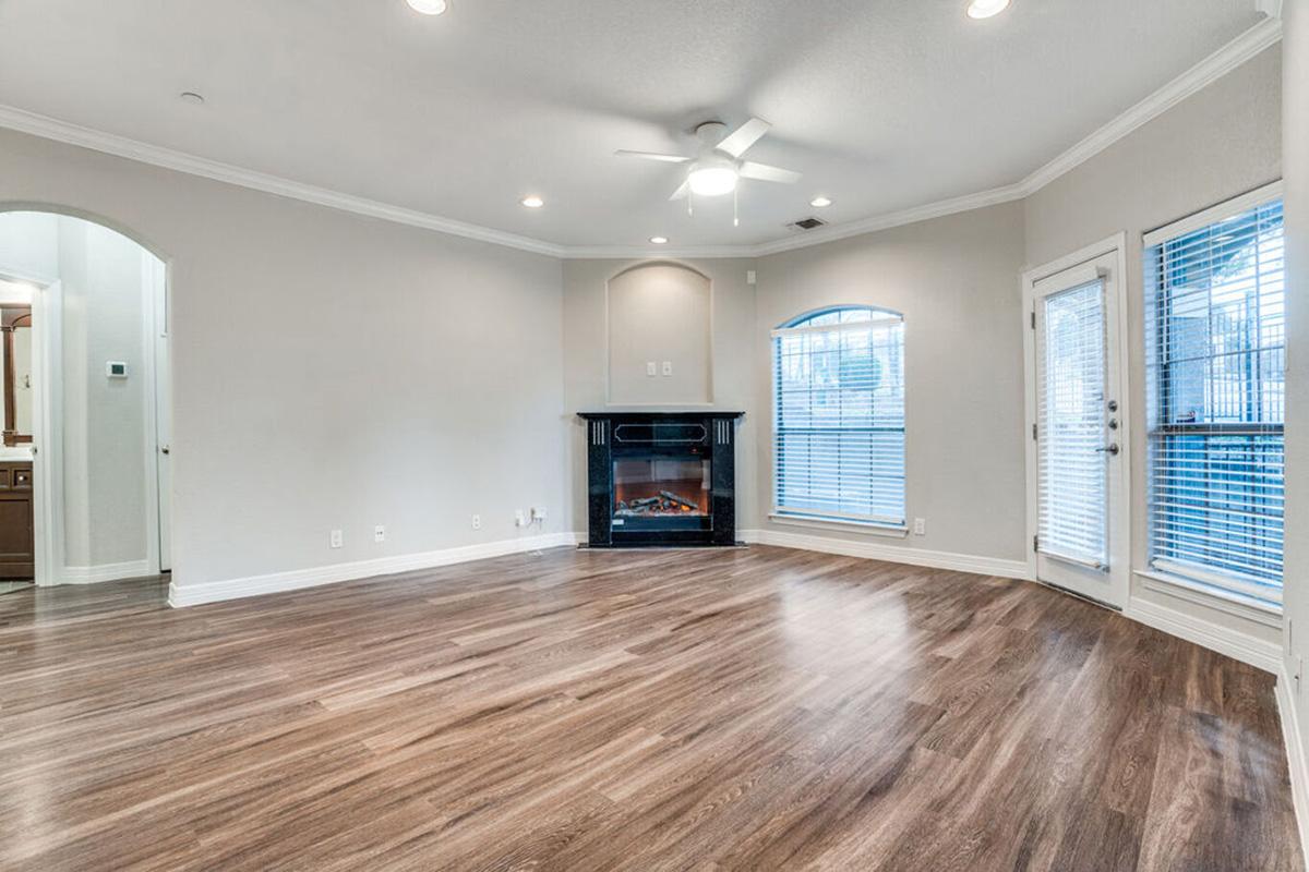Spacious living room with light hardwood flooring, a modern electric fireplace, and large windows with blinds. The room features a ceiling fan and a neutral color scheme, creating a bright and inviting atmosphere. There are two doorways leading to other areas of the home.