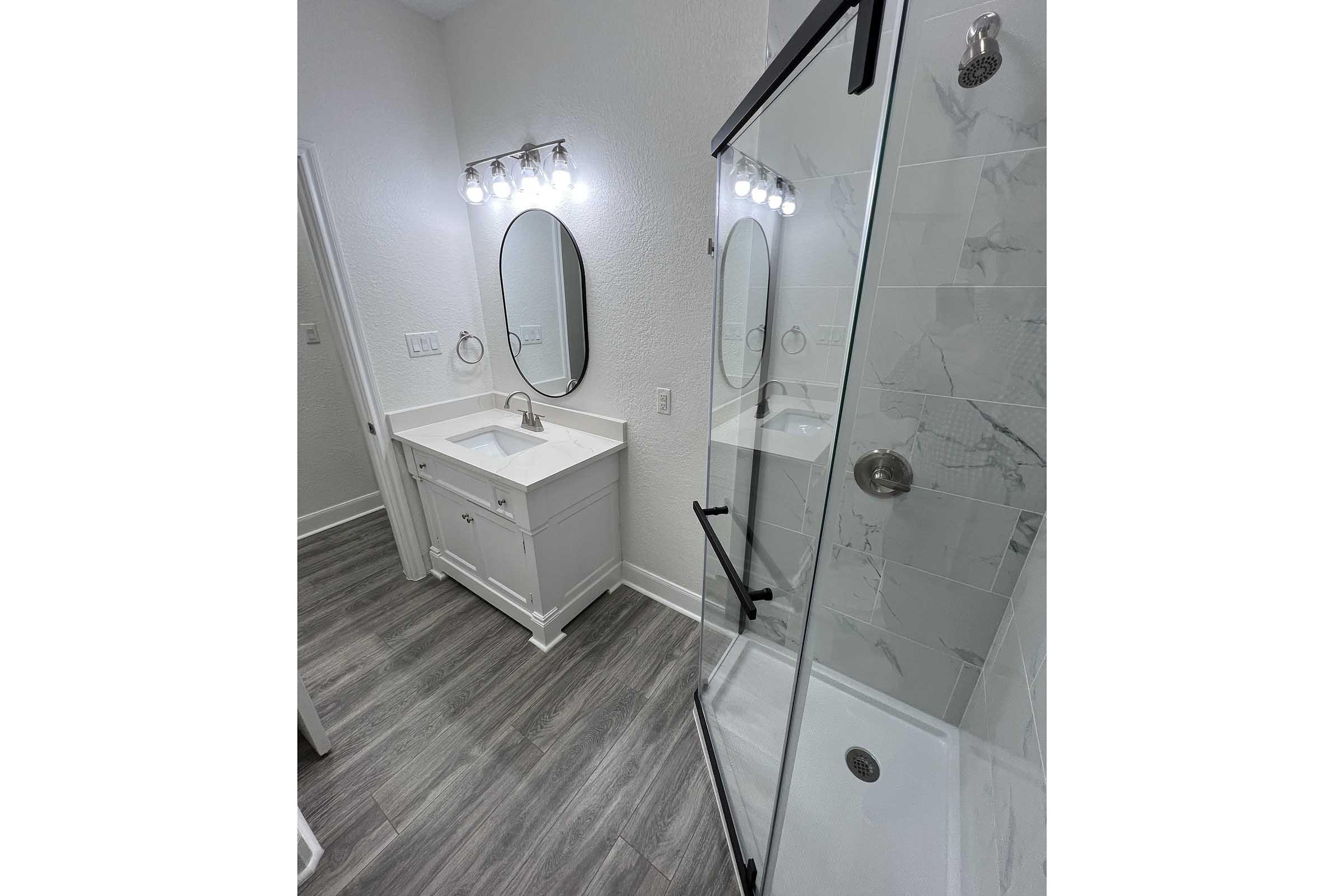 A well-lit, modern bathroom featuring a white vanity with a round mirror, a glass shower enclosure, and gray wood-like flooring. The shower has a sleek design with a glass door, and a row of light fixtures illuminates the space above the vanity.