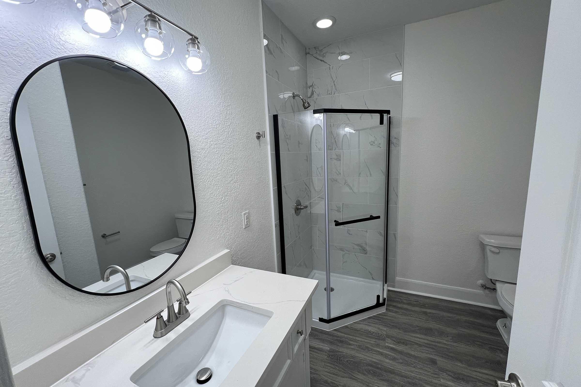 A modern bathroom featuring a circular mirror above a white vanity with a sink, a glass shower enclosure with black trim, a white toilet, and light-colored walls. The space has bright lighting and a sleek, contemporary design with gray flooring.