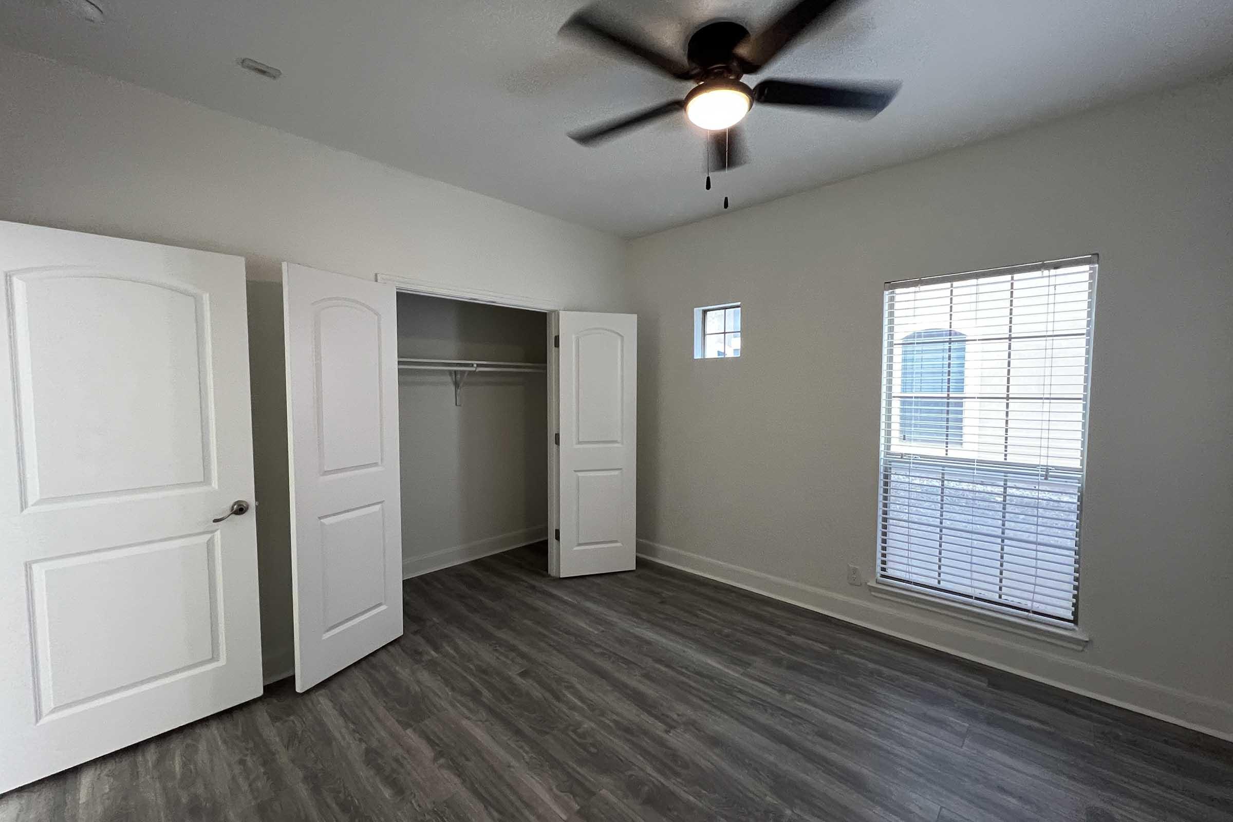 A bright, empty bedroom featuring light gray walls, a ceiling fan, and a large window with blinds. There are two white closet doors leading to a closet space. The floor has a wood-like laminate finish, contributing to a modern aesthetic in the room.