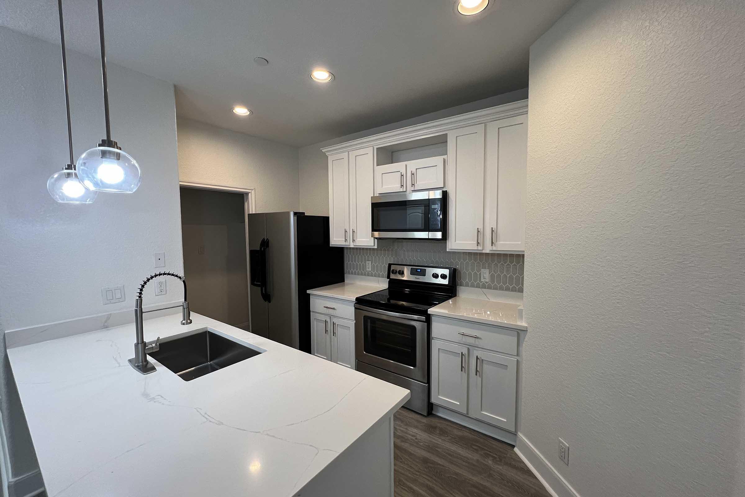 Modern kitchen featuring white cabinetry, stainless steel appliances, and a large island with a sink. The space has recessed lighting and a minimalist design with a neutral color palette. A sleek black refrigerator is visible, and the countertop has a marble-like finish.