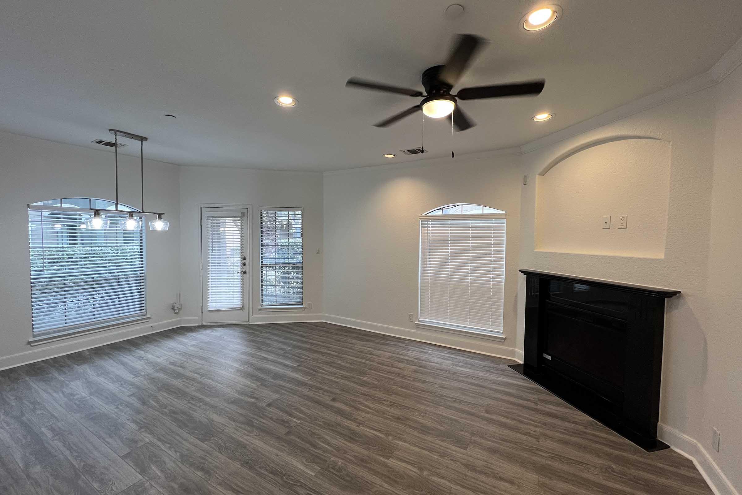 A spacious, well-lit living area featuring a ceiling fan, large windows with blinds, and a cozy fireplace. The flooring is a modern laminate, and there are sliding glass doors leading to a patio or outdoor space. The walls are painted in a neutral tone, creating a bright and inviting atmosphere.