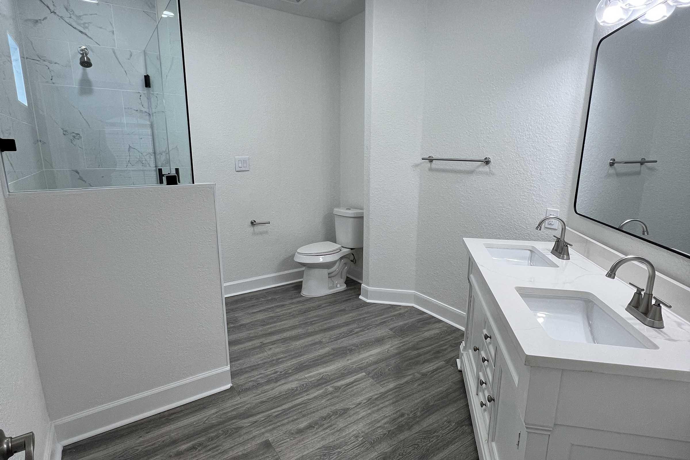 A modern bathroom featuring a glass shower enclosure, a white double vanity with two sinks, a toilet, and light wood plank flooring. The walls are painted white, and there is a large mirror above the vanity with three light fixtures.