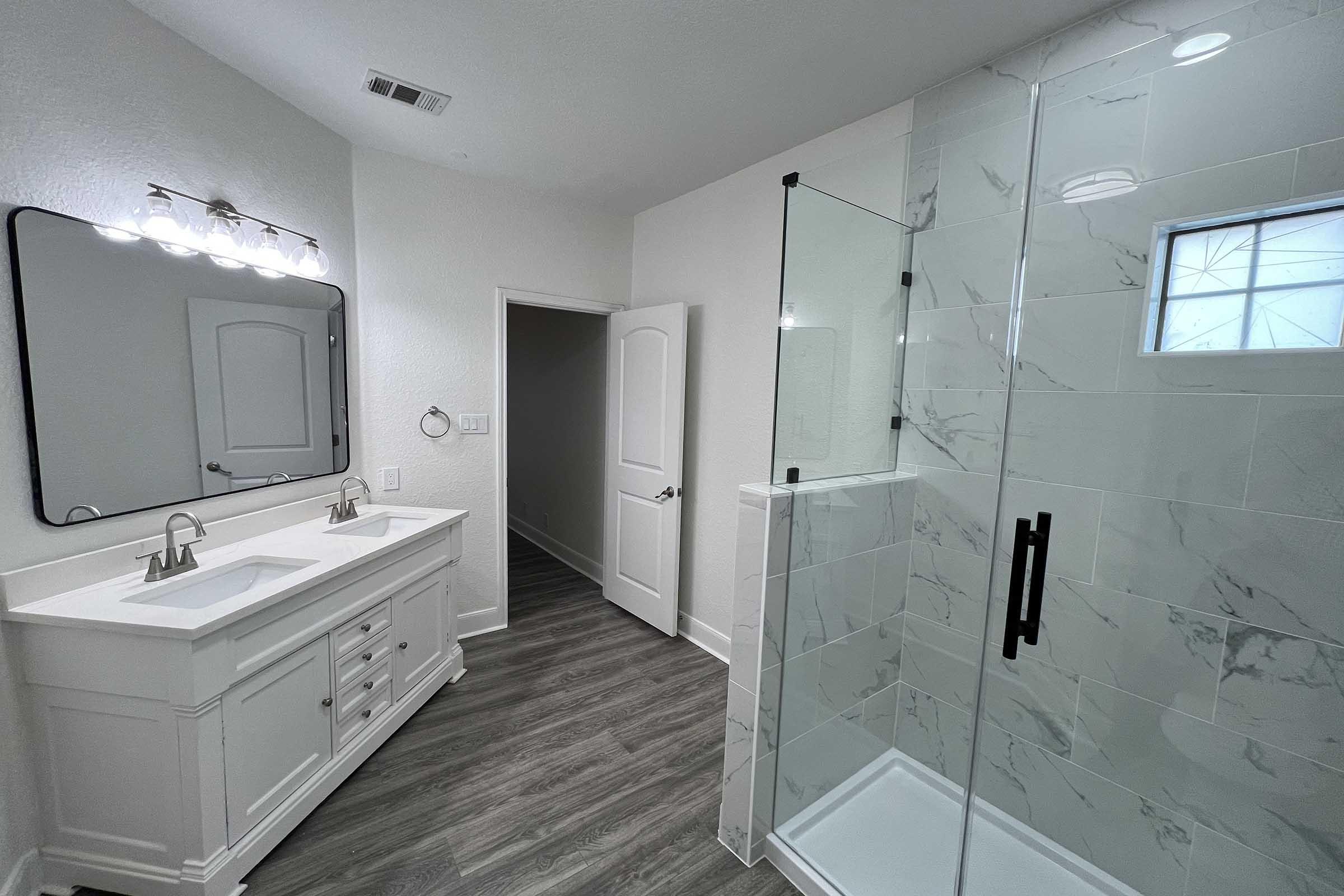 Modern bathroom featuring a double sink vanity with a large mirror, a walk-in shower with glass doors, and stylish marble tiles. The walls are painted in a light color, and the floor has a wood-like finish. A door leads to another room, adding a spacious feel to the layout.