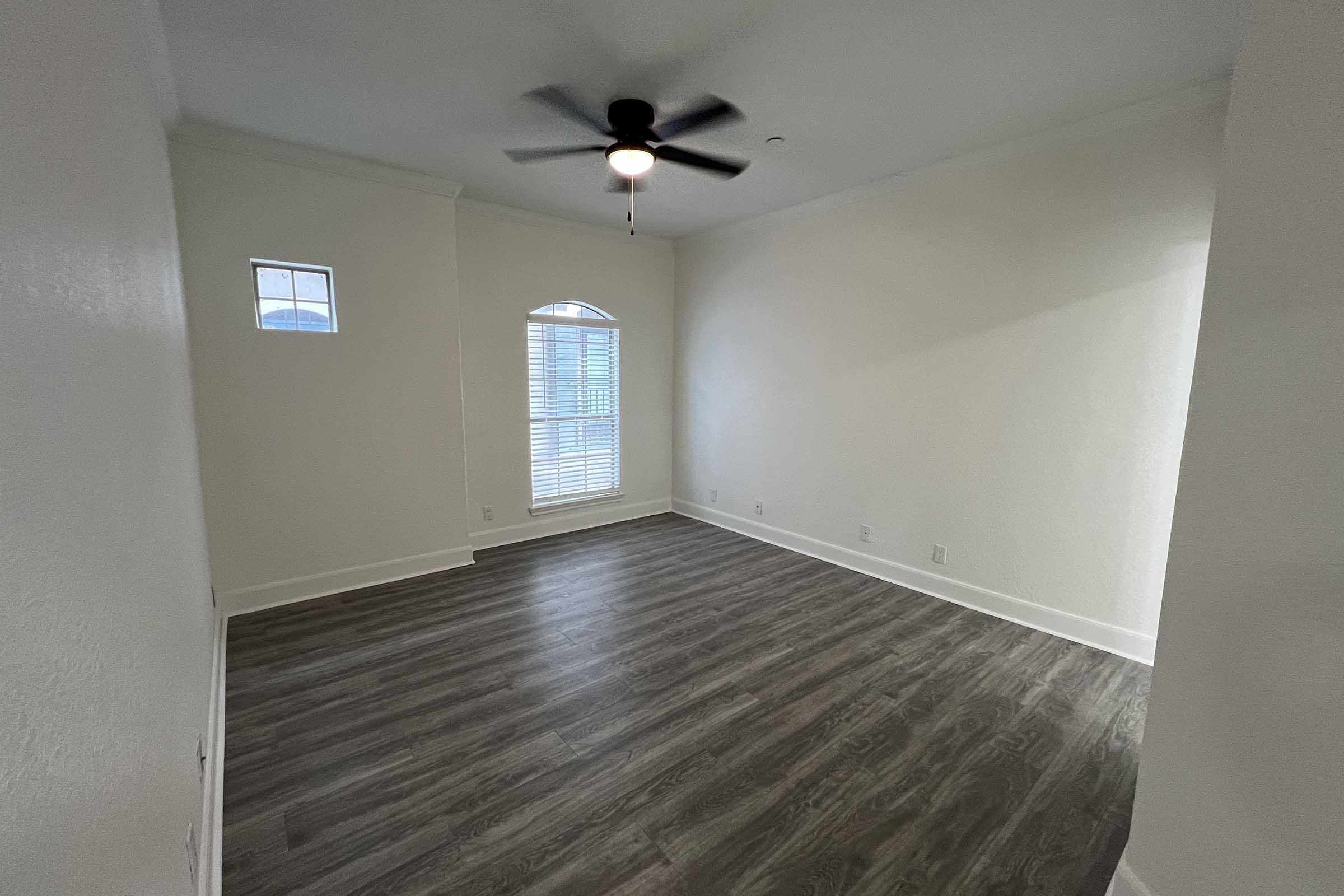 A spacious, empty room with light-colored walls and dark wood-like flooring. It features a ceiling fan, a small window, and an arched window allowing natural light. The room is well-lit and ready for furniture arrangement, creating a welcoming space.