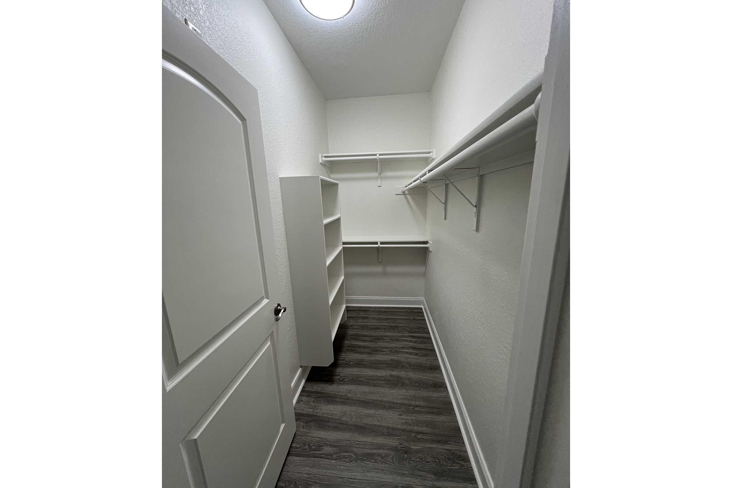 A small, empty closet featuring white walls, a ceiling light, and a dark wood floor. On the left, there is a white shelving unit, and along the back wall, there are two shelves mounted above the floor. The closet has a white door partially visible on the right side.