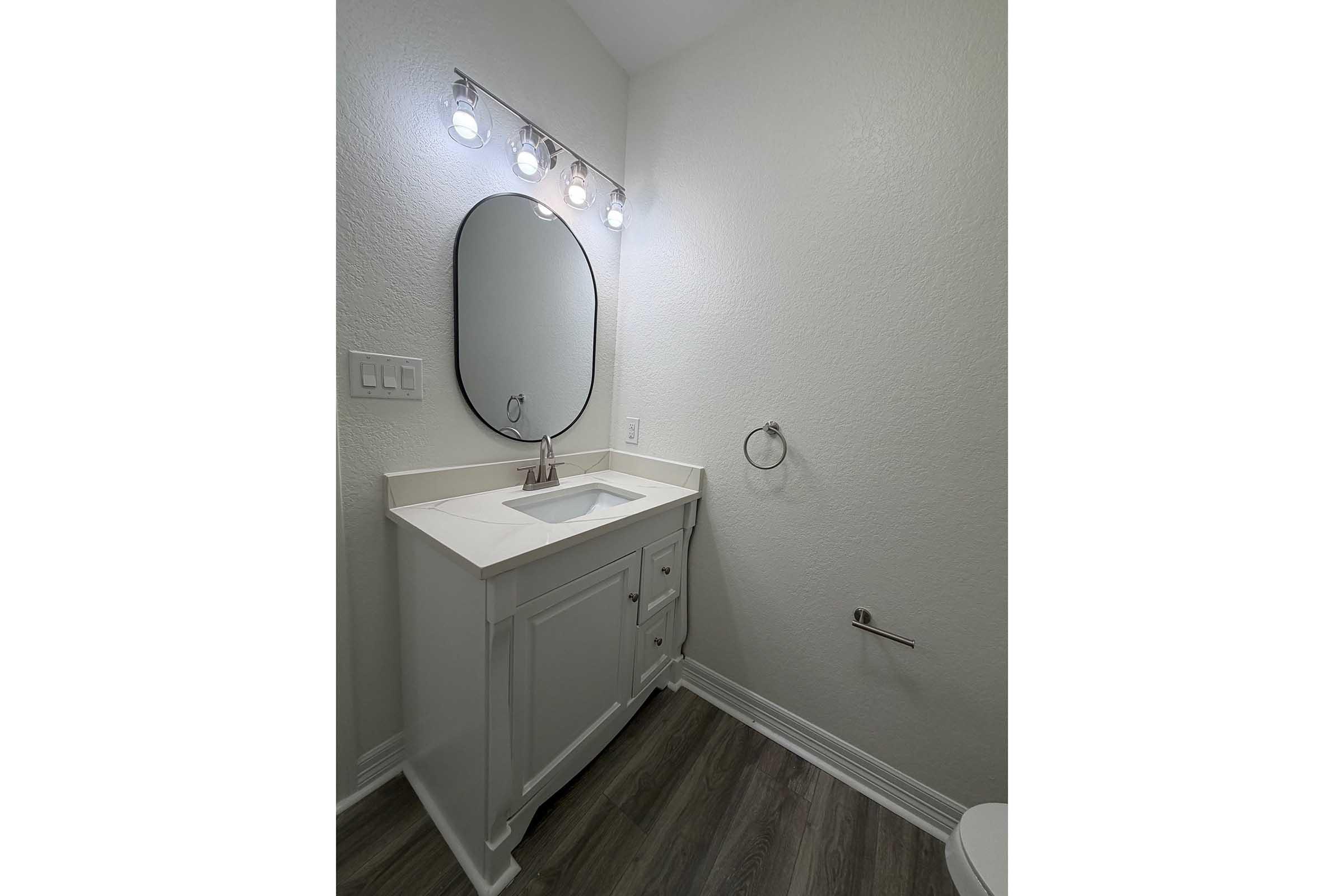 A modern bathroom featuring a white vanity with a sink, a large round mirror above it, and a stylish light fixture with four bulbs. The walls are painted light, and the flooring is made of wood-like material. A towel holder is mounted on the wall, and a toilet is visible in the corner.