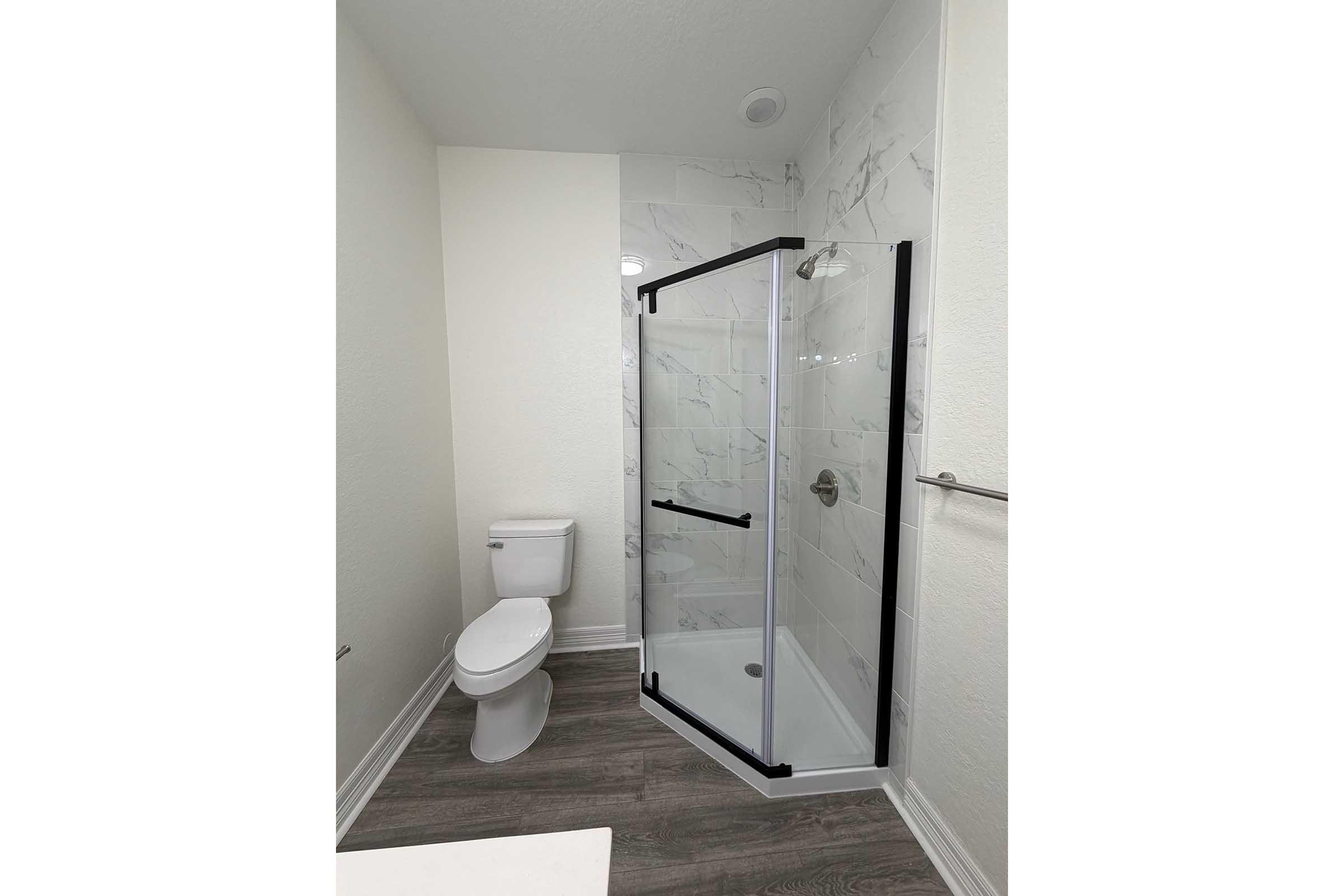 A modern bathroom featuring a glass shower enclosure with black accents, a white toilet, and light-colored walls. The shower area has a marble-like appearance, and there is a towel rack on the wall. The flooring is a wood-like laminate, creating a clean and contemporary aesthetic.