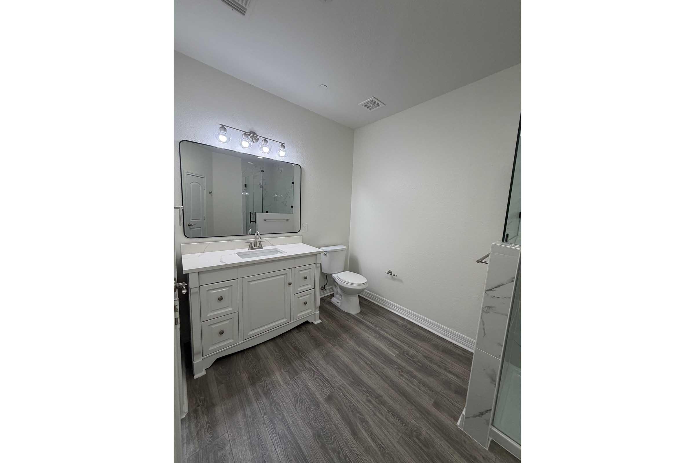 A modern bathroom featuring a light-colored vanity with a sink and a large mirror above, a white toilet, and a glass-enclosed shower. The walls are painted a neutral tone, and the floor is covered with brown wood-like laminate. Bright lighting is provided by fixtures above the mirror.