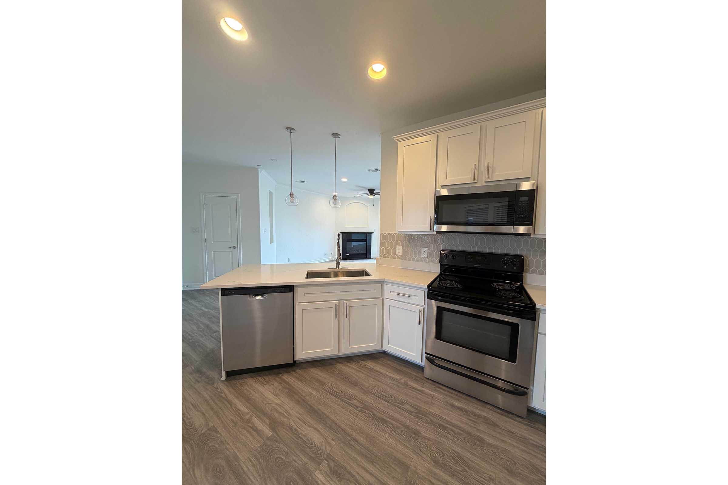 Modern kitchen featuring white cabinetry, a stainless steel dishwasher, and a black oven range. Quartz countertop with a sink and pendant lighting above. The open layout leads to a living area with light-colored walls and hardwood-style flooring. Natural light brightens the space.