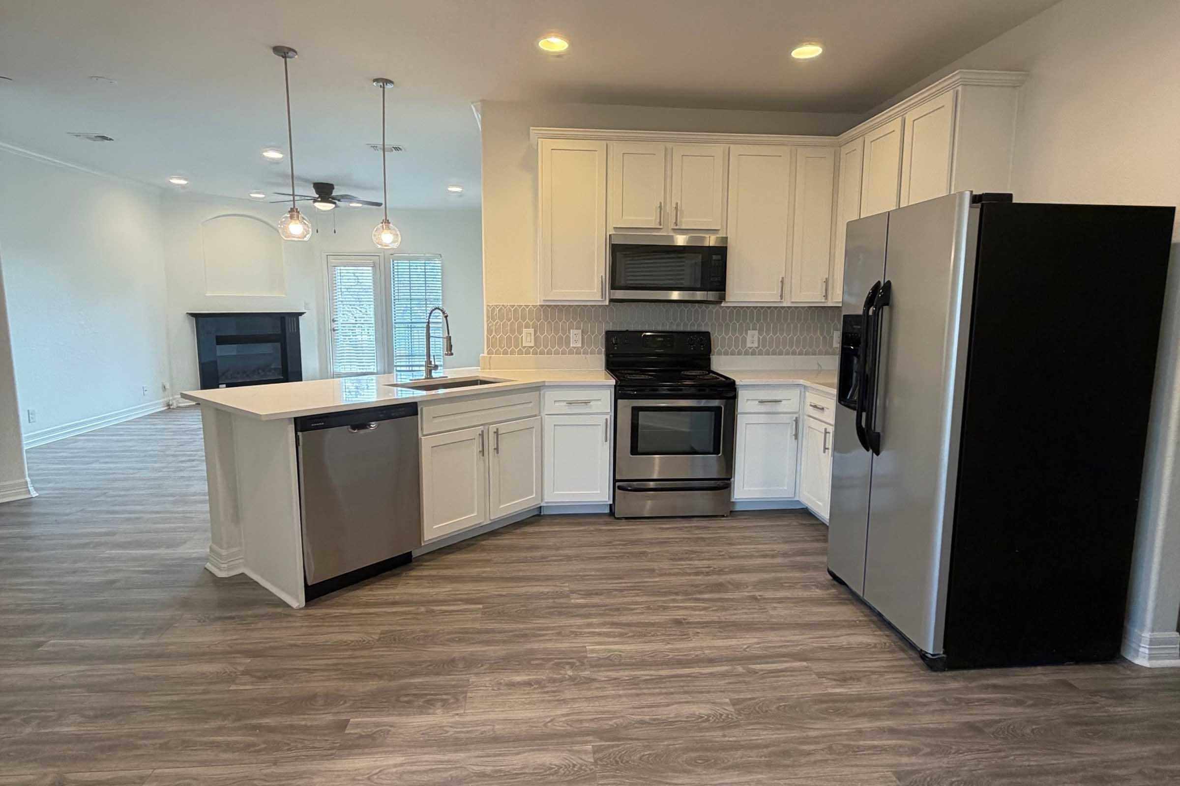 Modern kitchen featuring white cabinetry, stainless steel appliances including an oven, microwave, and dishwasher, and a black refrigerator. The space has a peninsula with seating and is set against light-colored walls and wood-look flooring, with a view into a living area with a fireplace.