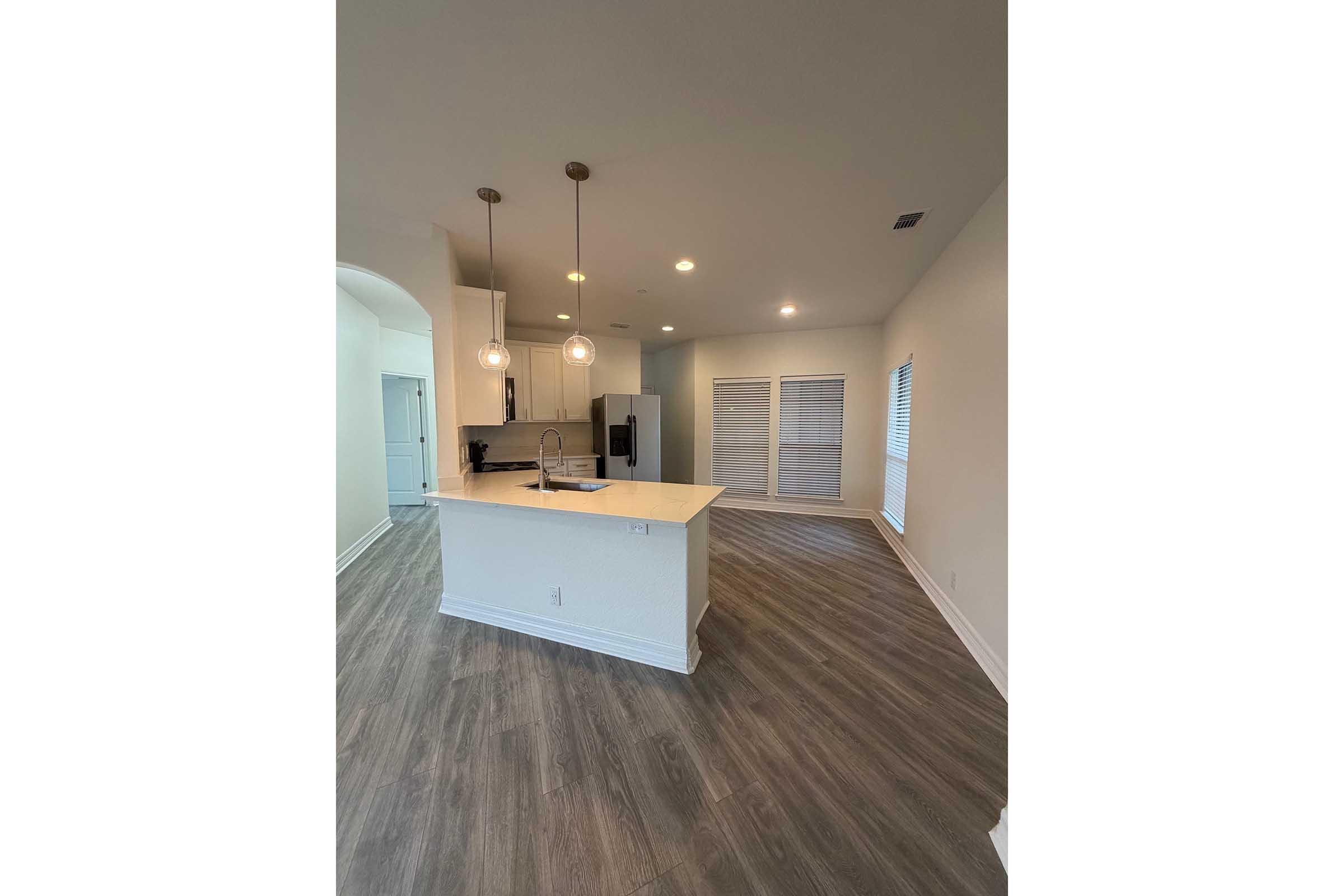 Modern kitchen featuring a central island with a sink and pendant lighting, white cabinetry, stainless steel appliances, and a spacious open layout. Large windows provide natural light, complementing the wood-look flooring and neutral wall colors.